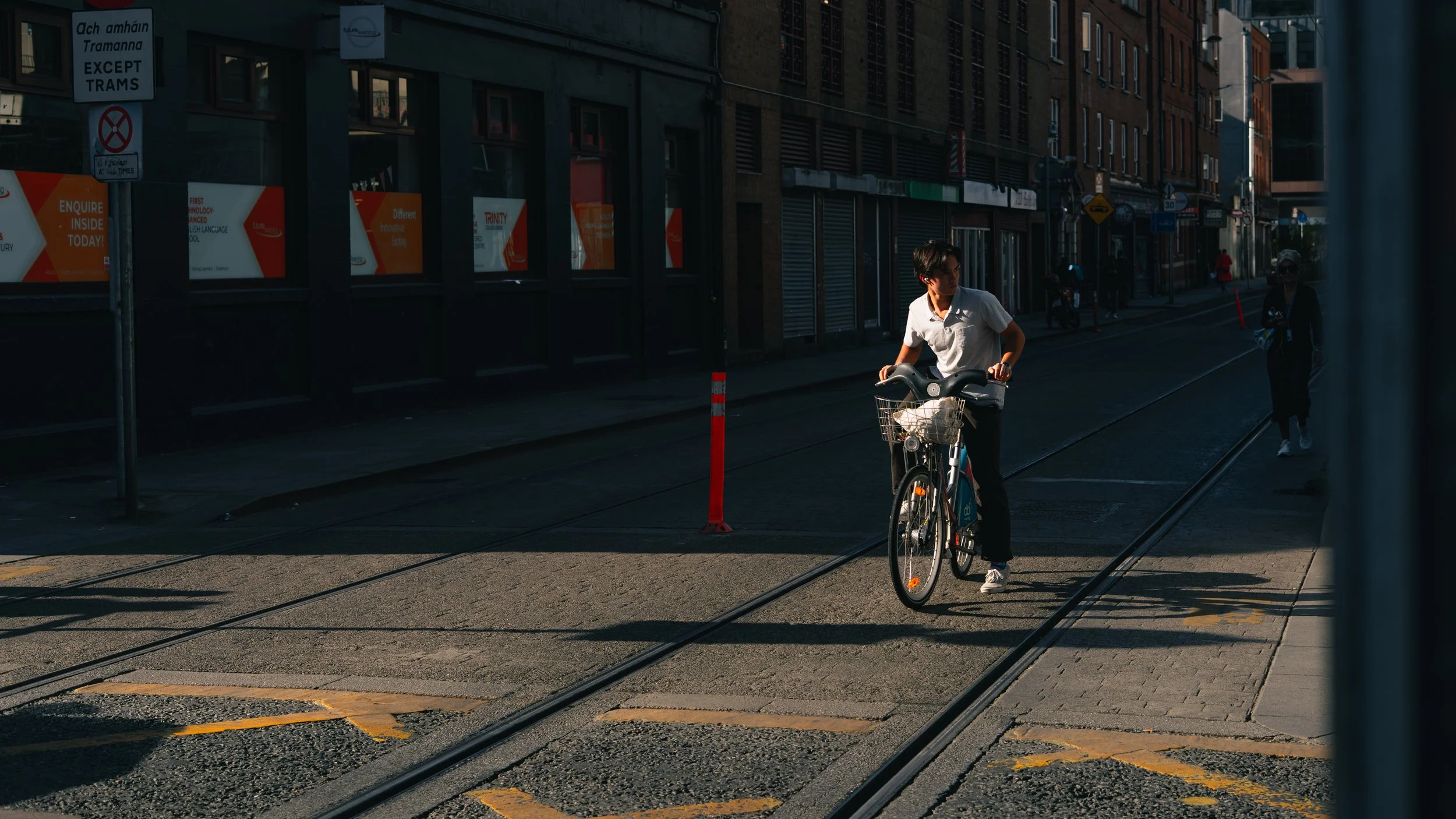 A person in a white shirt is riding a bicycle across a street with tram tracks. The street is partially in shadow, and there are a few other pedestrians walking along the sidewalk in the background.