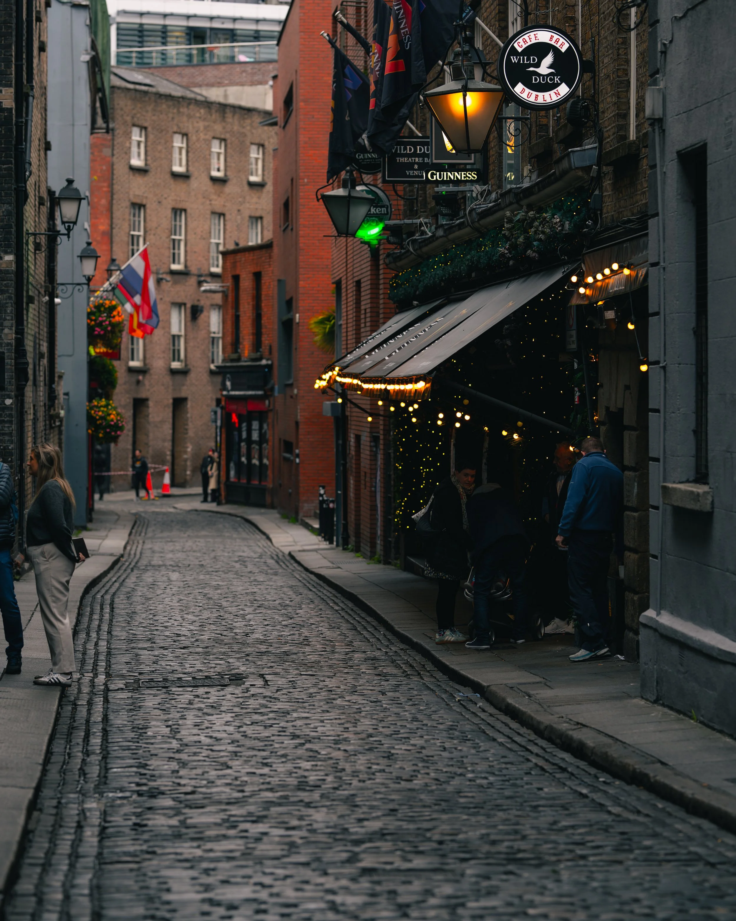 A narrow cobblestone street in Dublin with pedestrians, illuminated signs, and a pub with string lights outside.