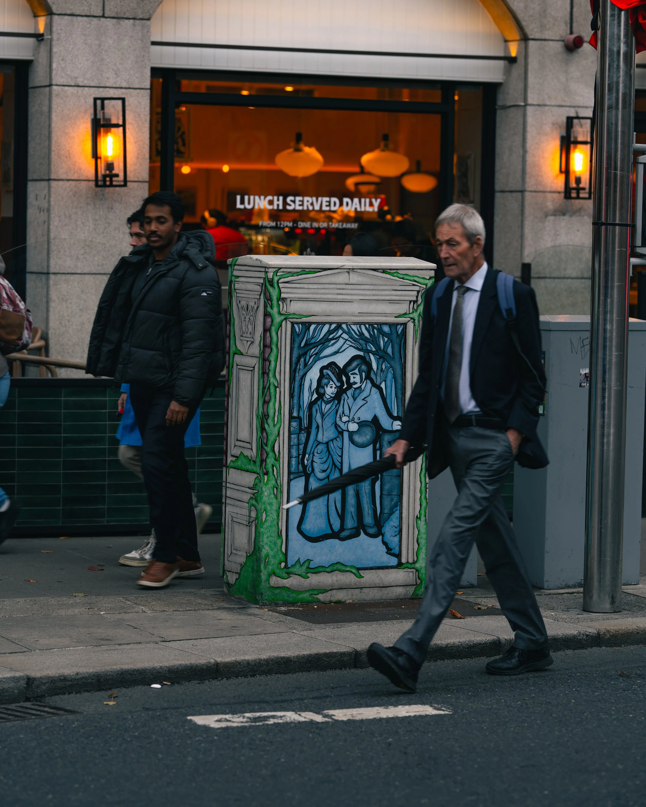 People walking on city sidewalk in front of a colorful painted utility box with a historical scene. A man in a suit with an umbrella walks past, while others pass by, with a restaurant in the background advertising daily lunch.