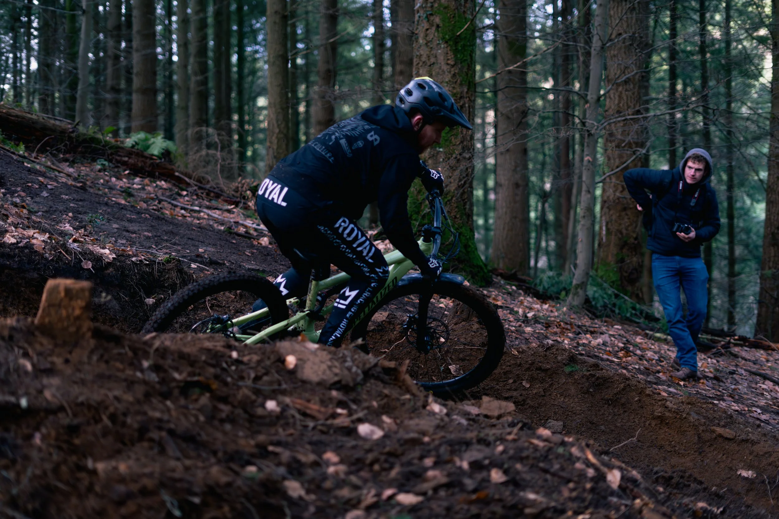 A person riding a mountain bike downhill on a forest trail, while another person stands nearby observing, holding a camera.
