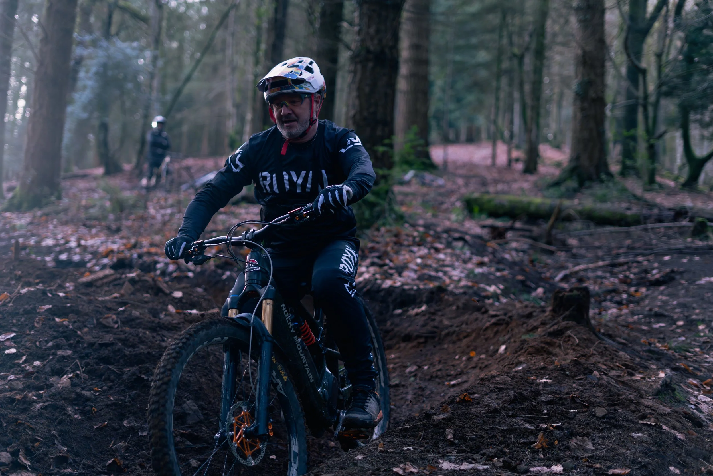 A man mountain biking on a trail in a forest with another cyclist in the background.