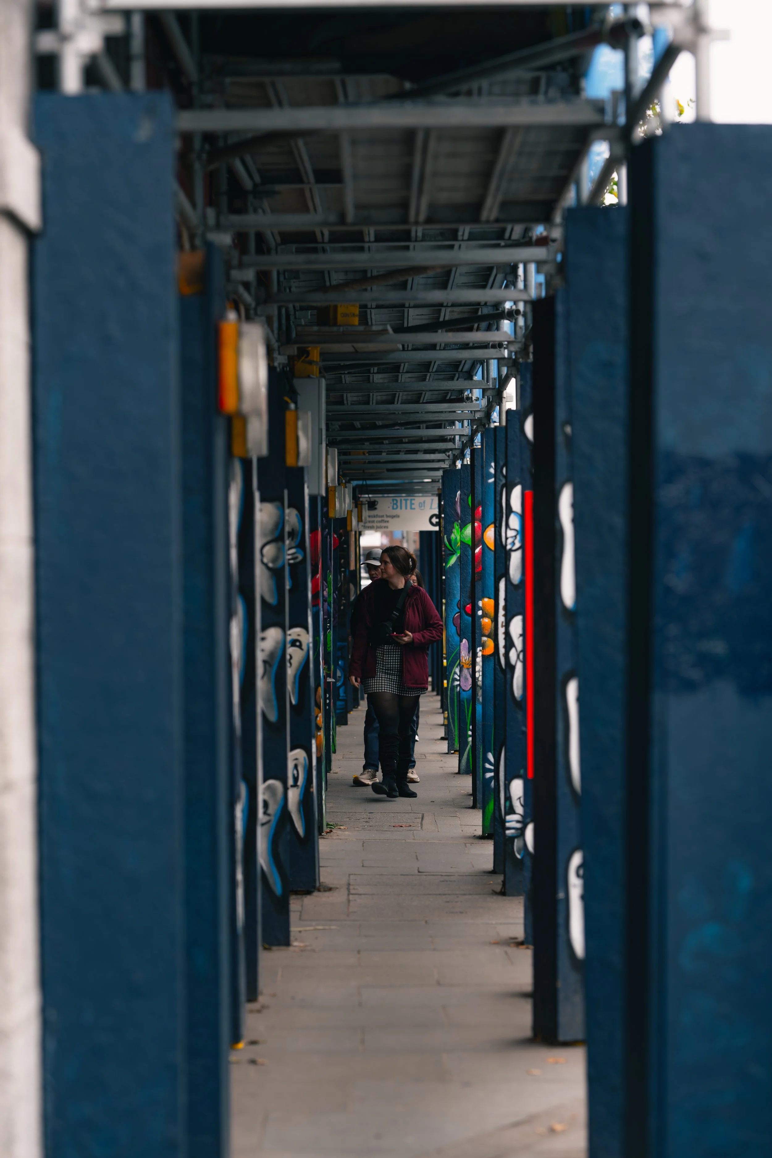 People walking through a narrow alleyway with blue painted walls and colorful graffiti art on the side panels during daytime.