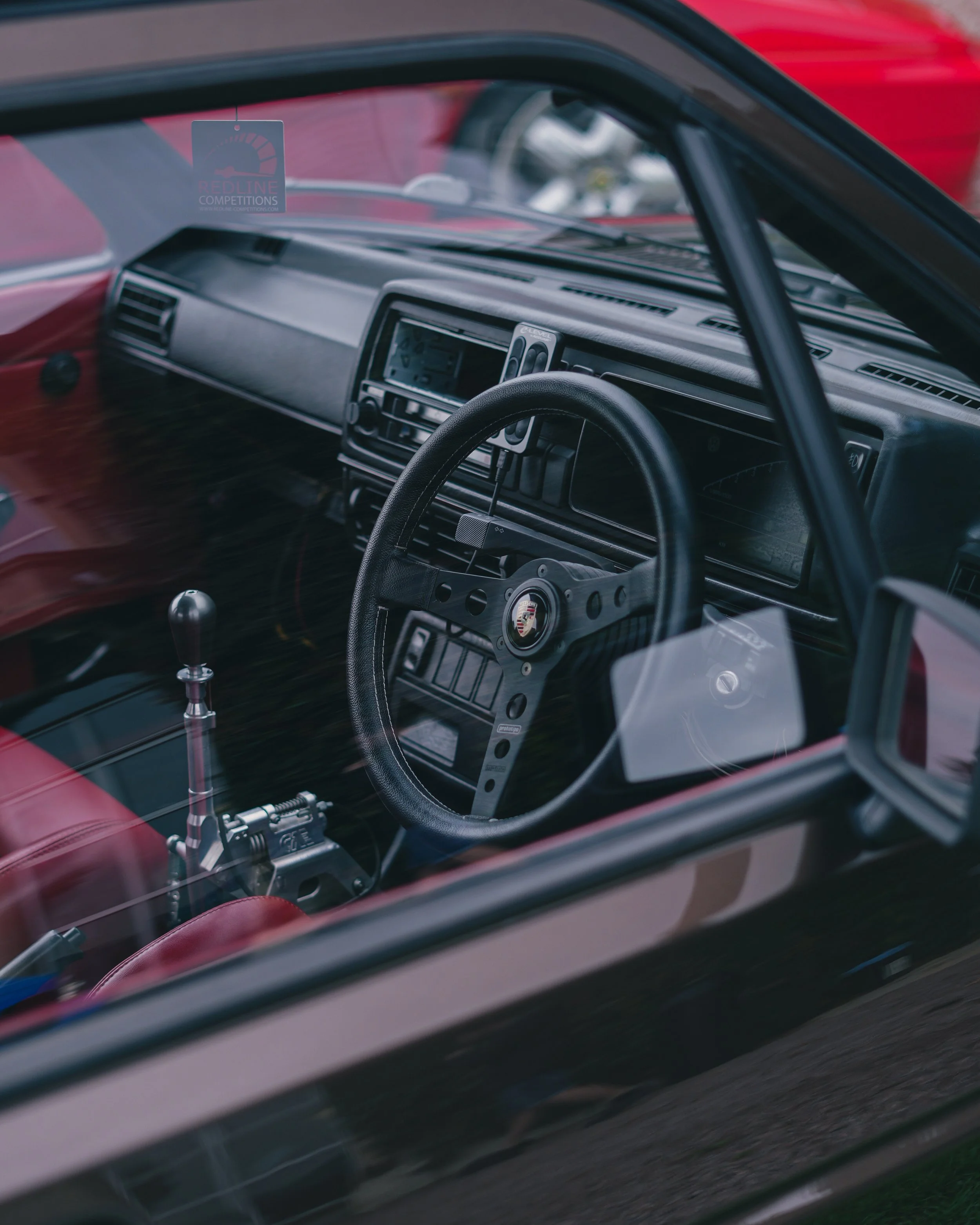 The interior of a vintage Volkswagen Golf car with a Porsche logo steering wheel, a gear shifter, and a dashboard with various controls, seen through the windshield.