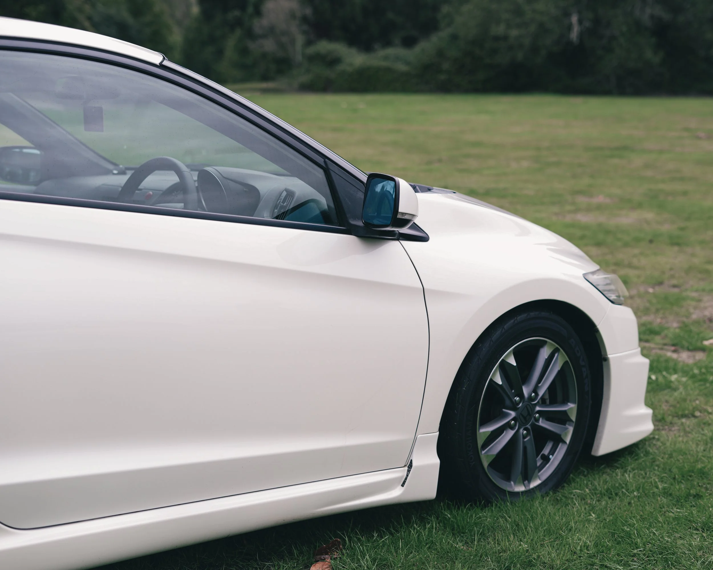 Close-up of a white Honda CR-Z sports car parked on a grassy field, showing the driver's side with a view of the side mirror and part of the tire.