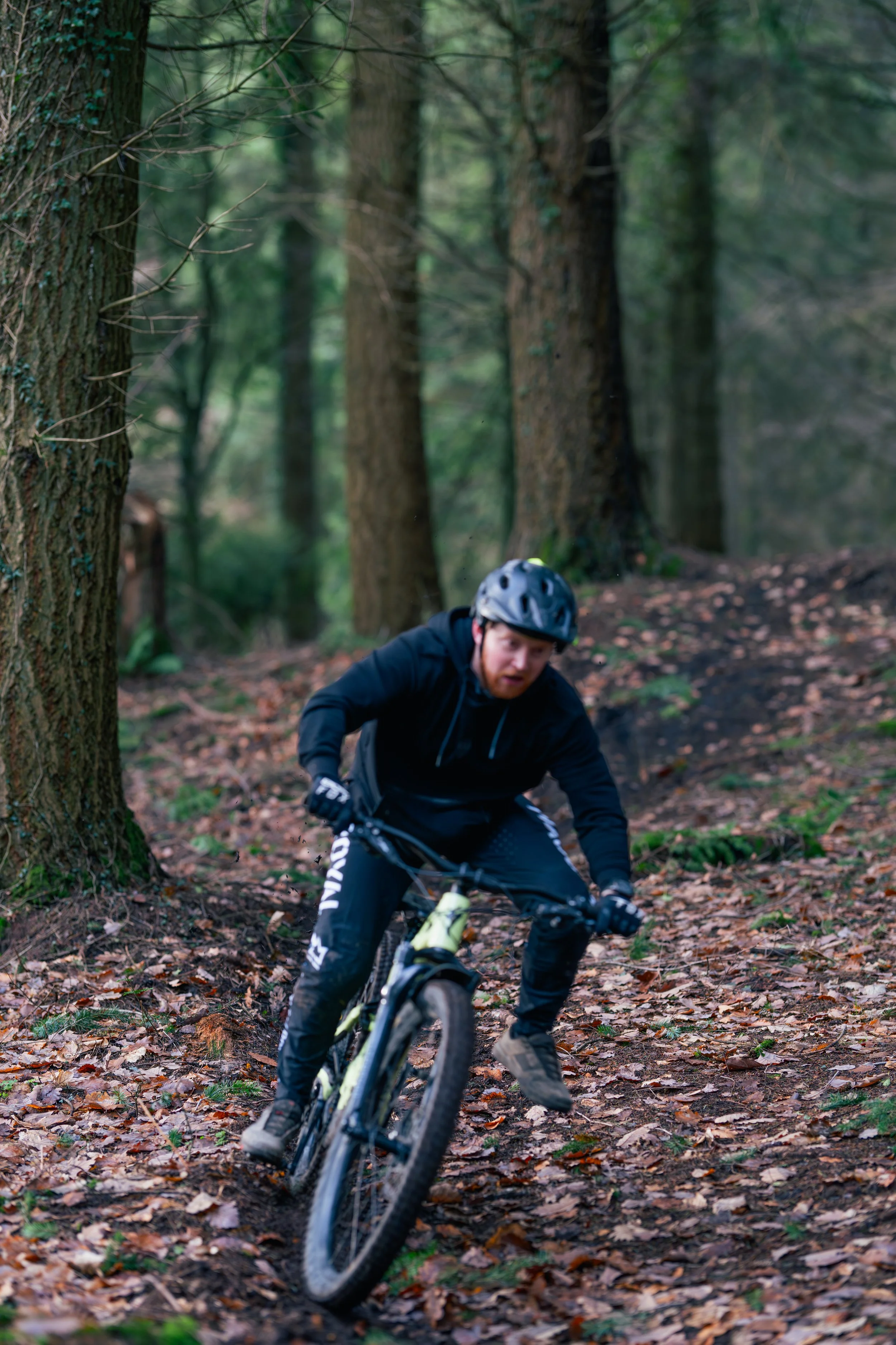 A man wearing a helmet and black athletic clothing riding a mountain bike on a trail covered in fallen leaves in a dense forest.