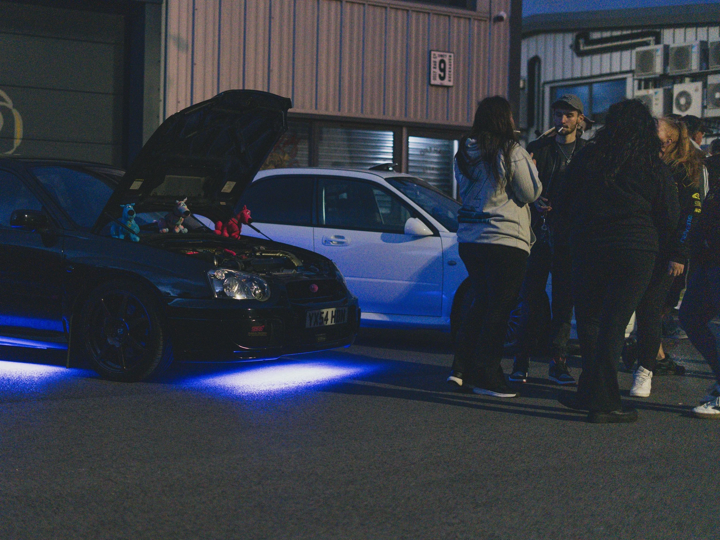 Group of people gathered outside at night near two cars, one with its hood open and underglow lights, with stuffed animals on the engine, next to a building with a sign.