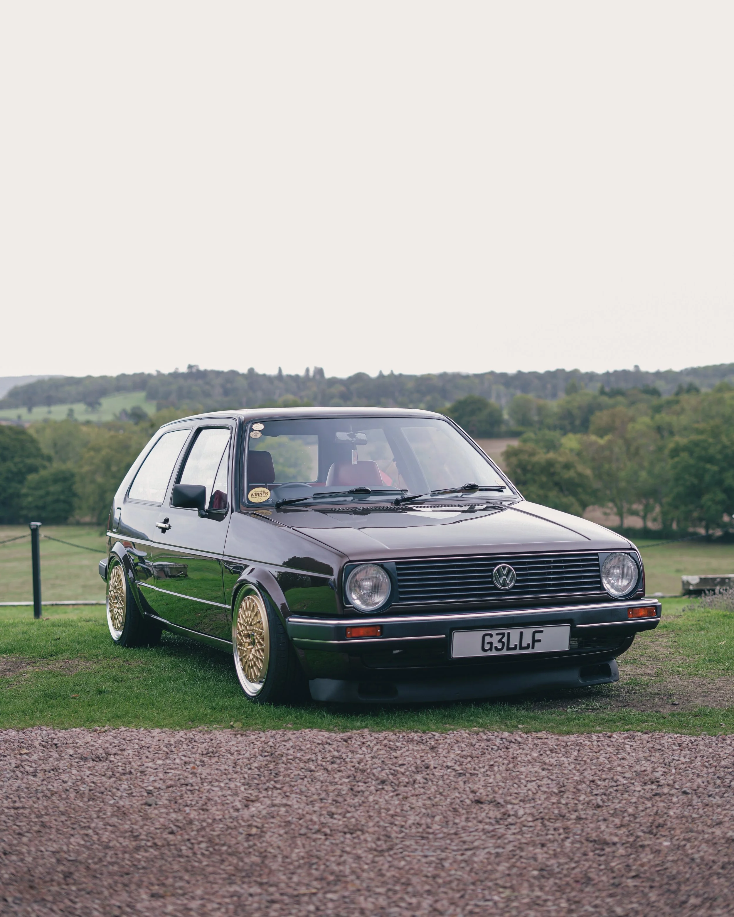 A vintage black Volkswagen Golf Mk2 parked on grass with a countryside background.