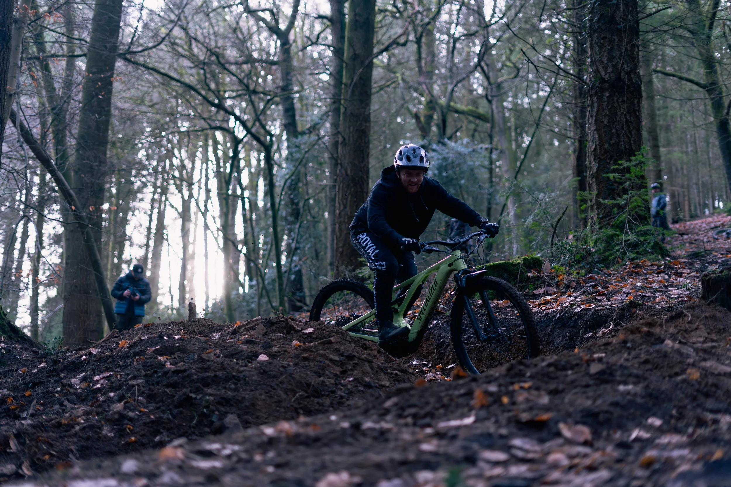 A person mountain biking on a trail in a forest with tall trees, wearing a helmet and black clothing, with two other people in the background taking photos.