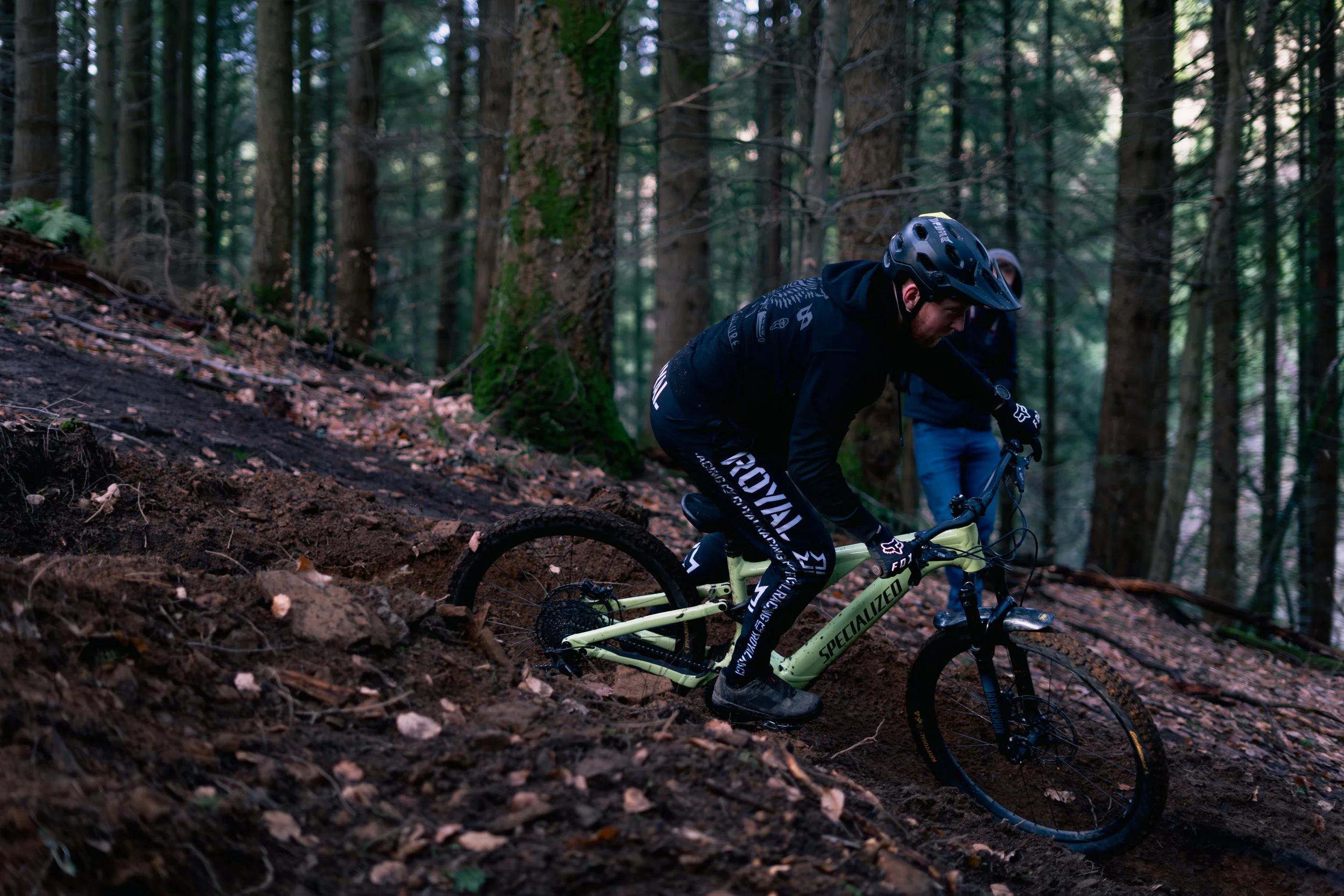 A mountain biker in black gear riding downhill on a dirt trail in a dense forest.