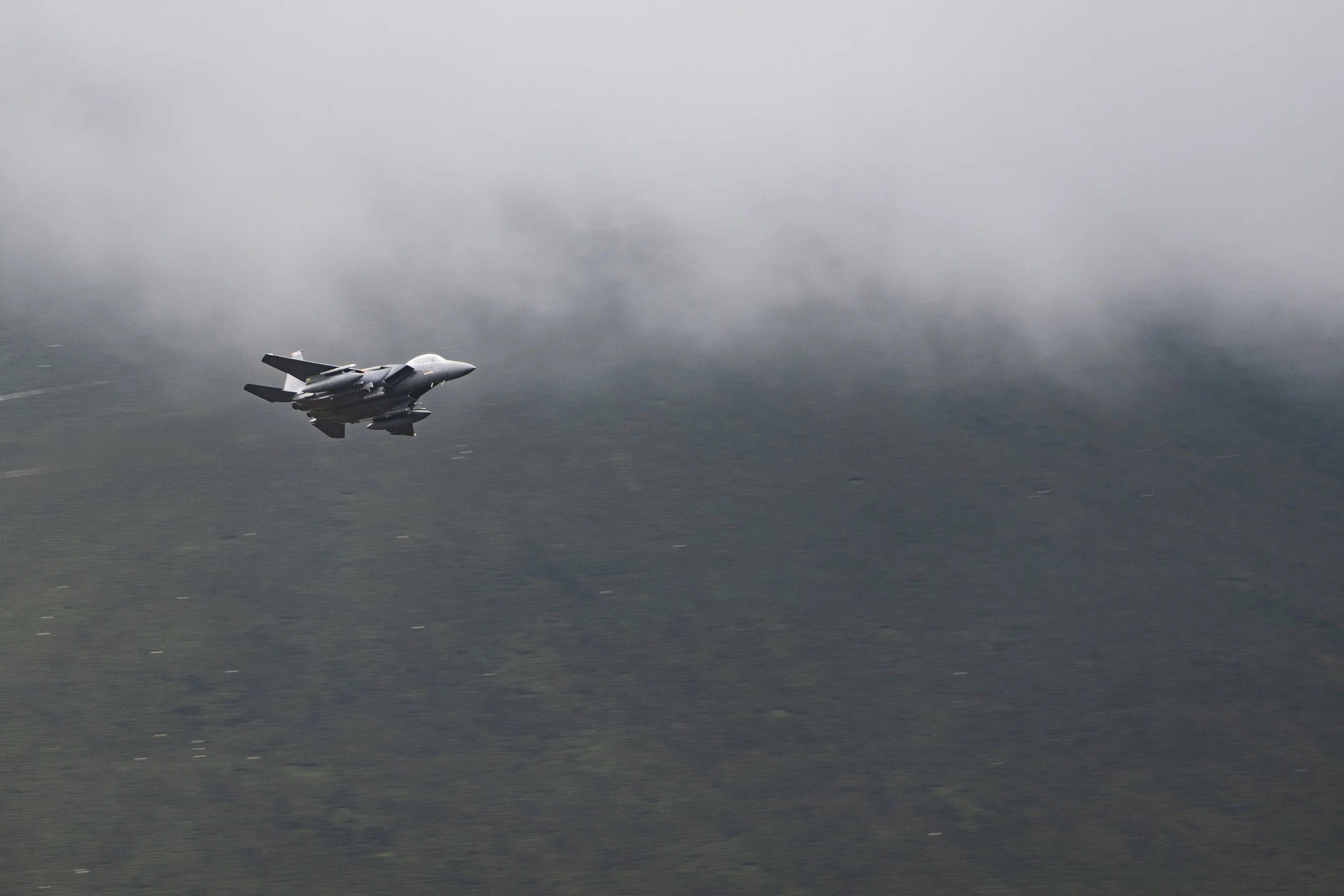 A fighter jet flying through cloudy skies over a forested area.