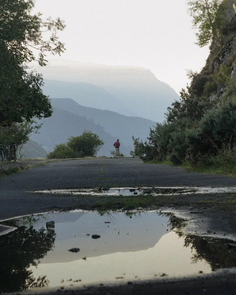 A person Infront of the Snowdonia national park on a mountain road with trees and bushes on both sides, overlooking distant layered mountains, with puddles on the road reflecting the sky and surroundings.