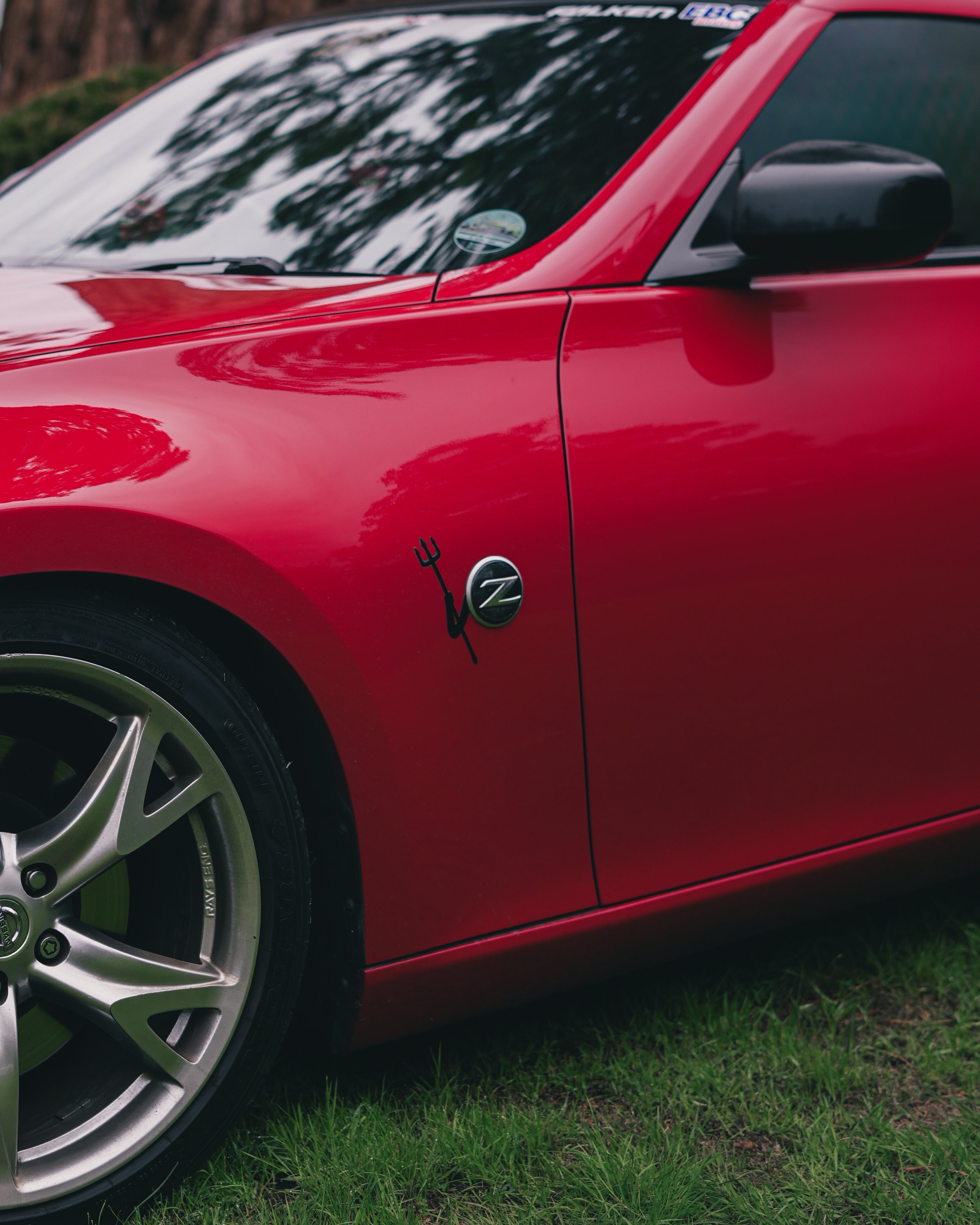 Close-up of a red  Nissan z370 sports car parked on grass, featuring a custom decal of a trident and a 'Z' emblem on the front fender, with reflections of trees on the windshield.