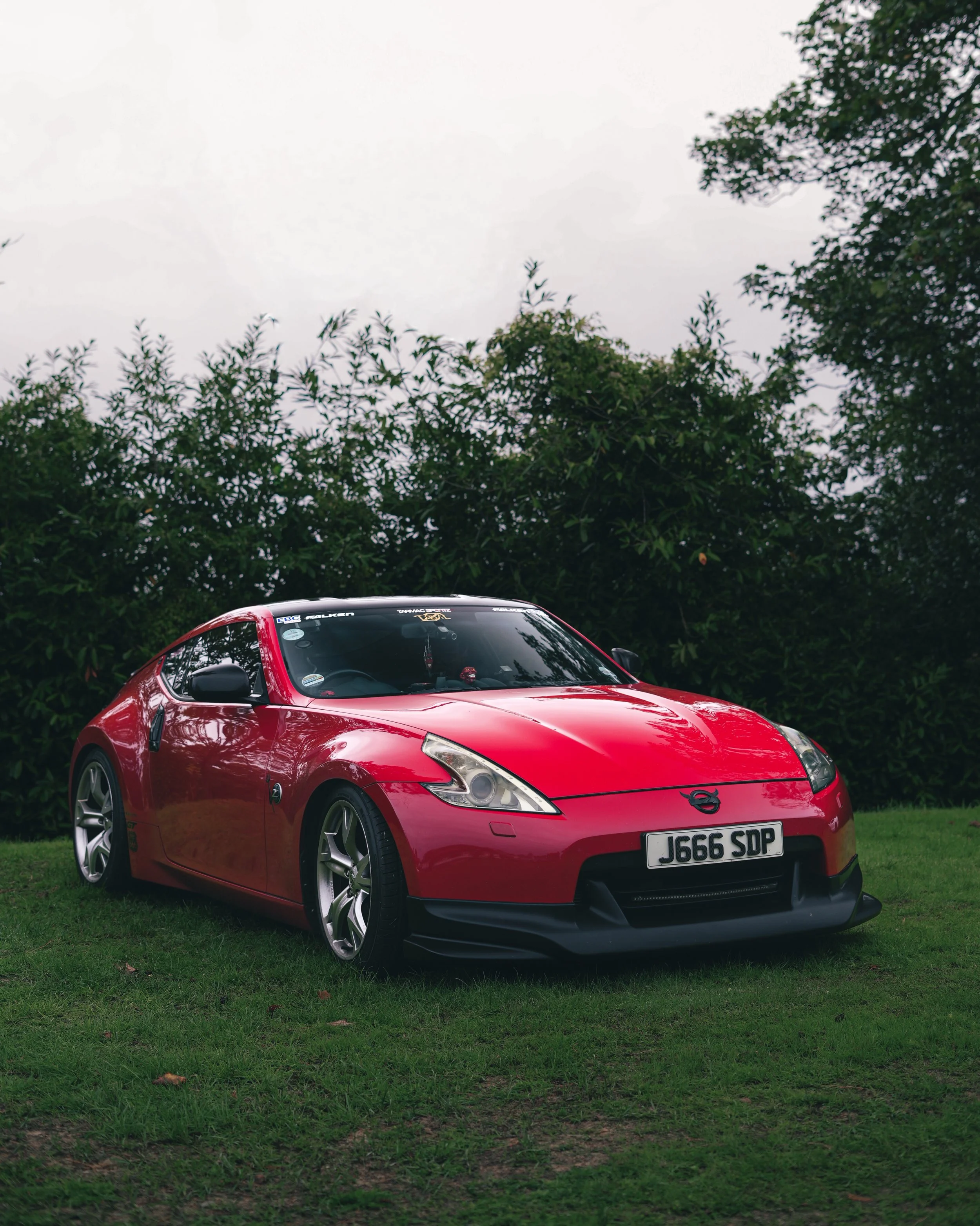 A red Nissan z370 sports car parked on a grassy area with trees in the background.
