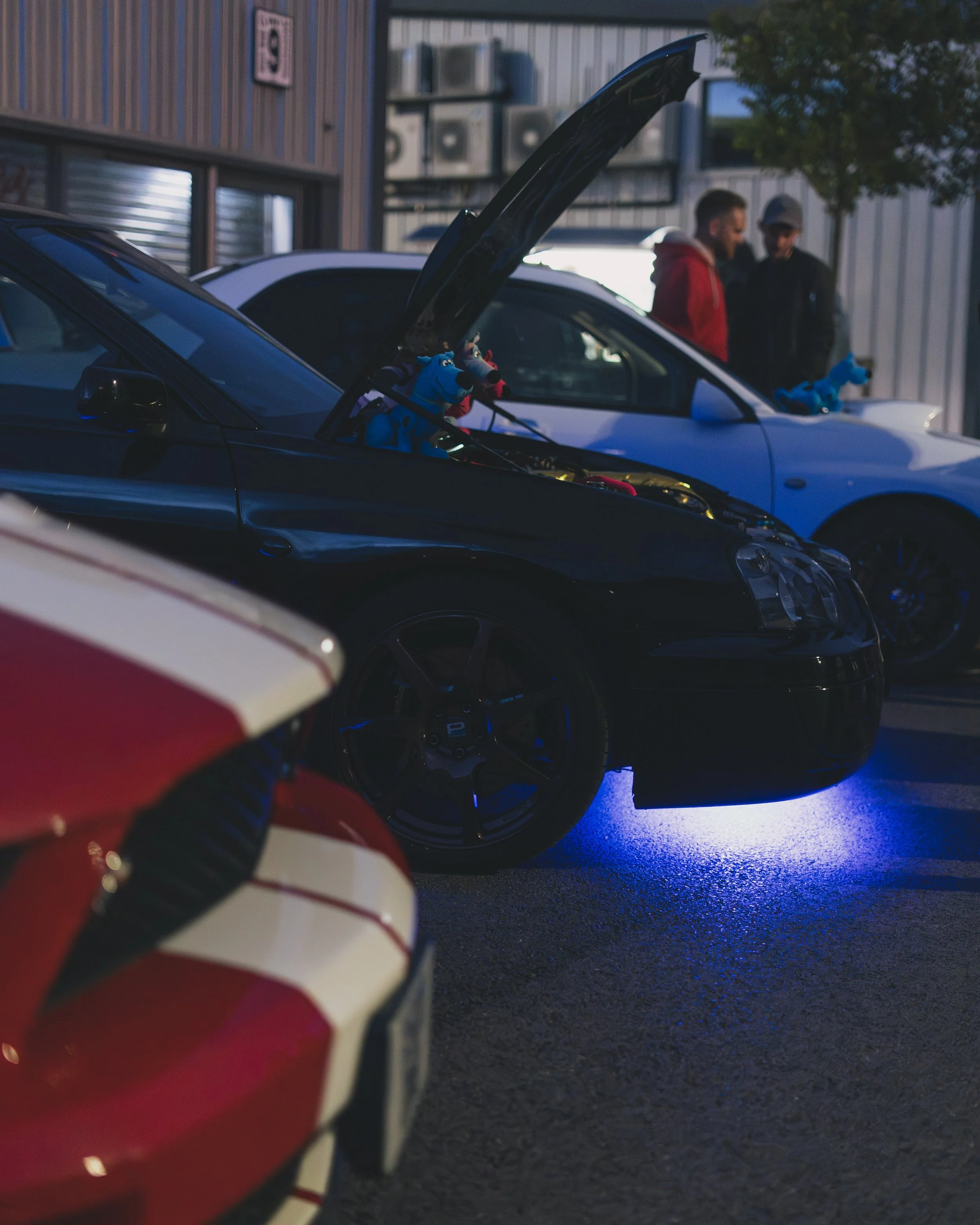 Black sports car with its hood open, showing engine, parked at night with blue underglow lights, with a red and white race car partially visible in the foreground, and two men talking near a white car in the background.