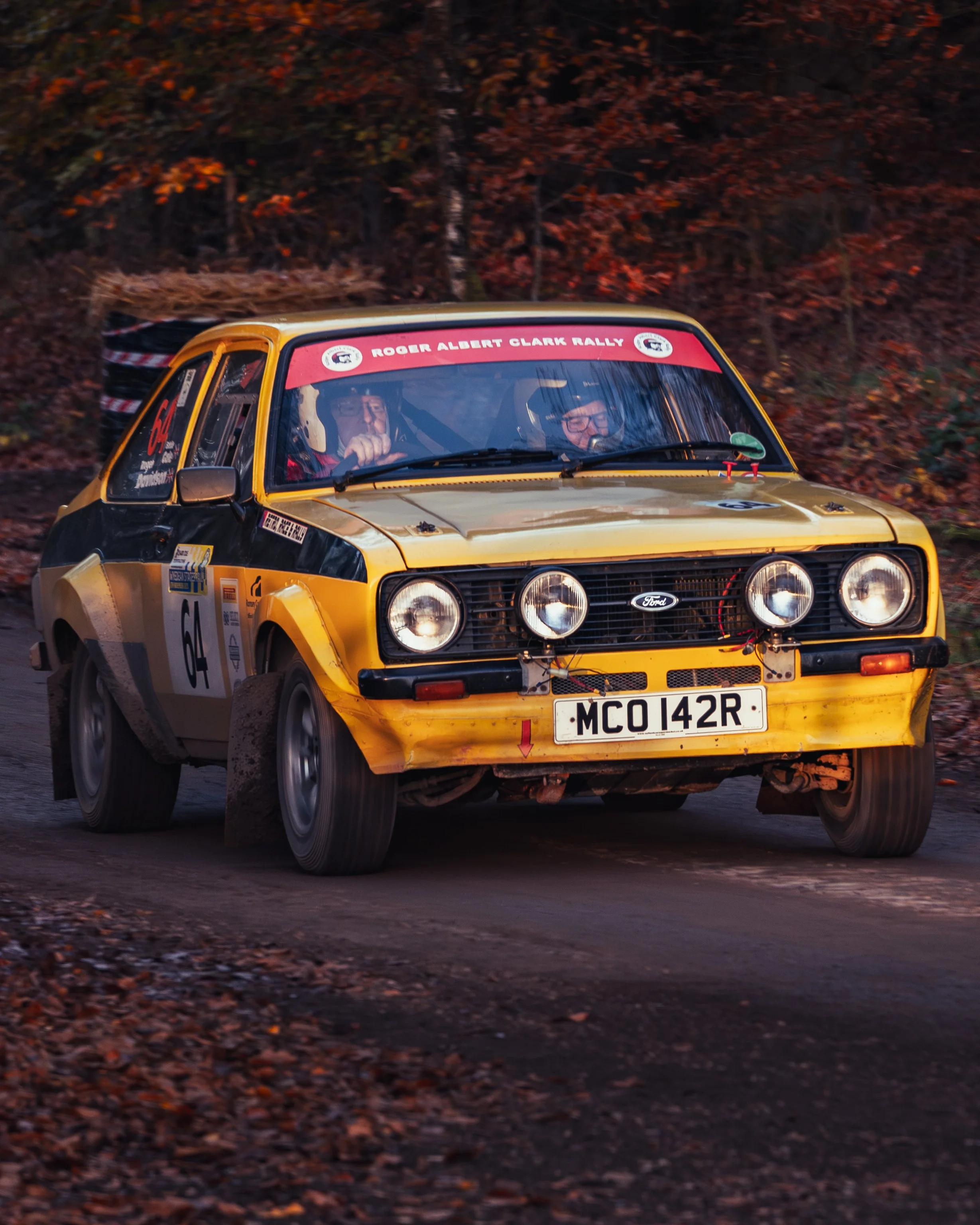 A yellow Ford rally car with four round headlights and a registration number MCO 142R driving on a dirt road, with a wooded forest background, during the Roger Albert Clark Rally. 