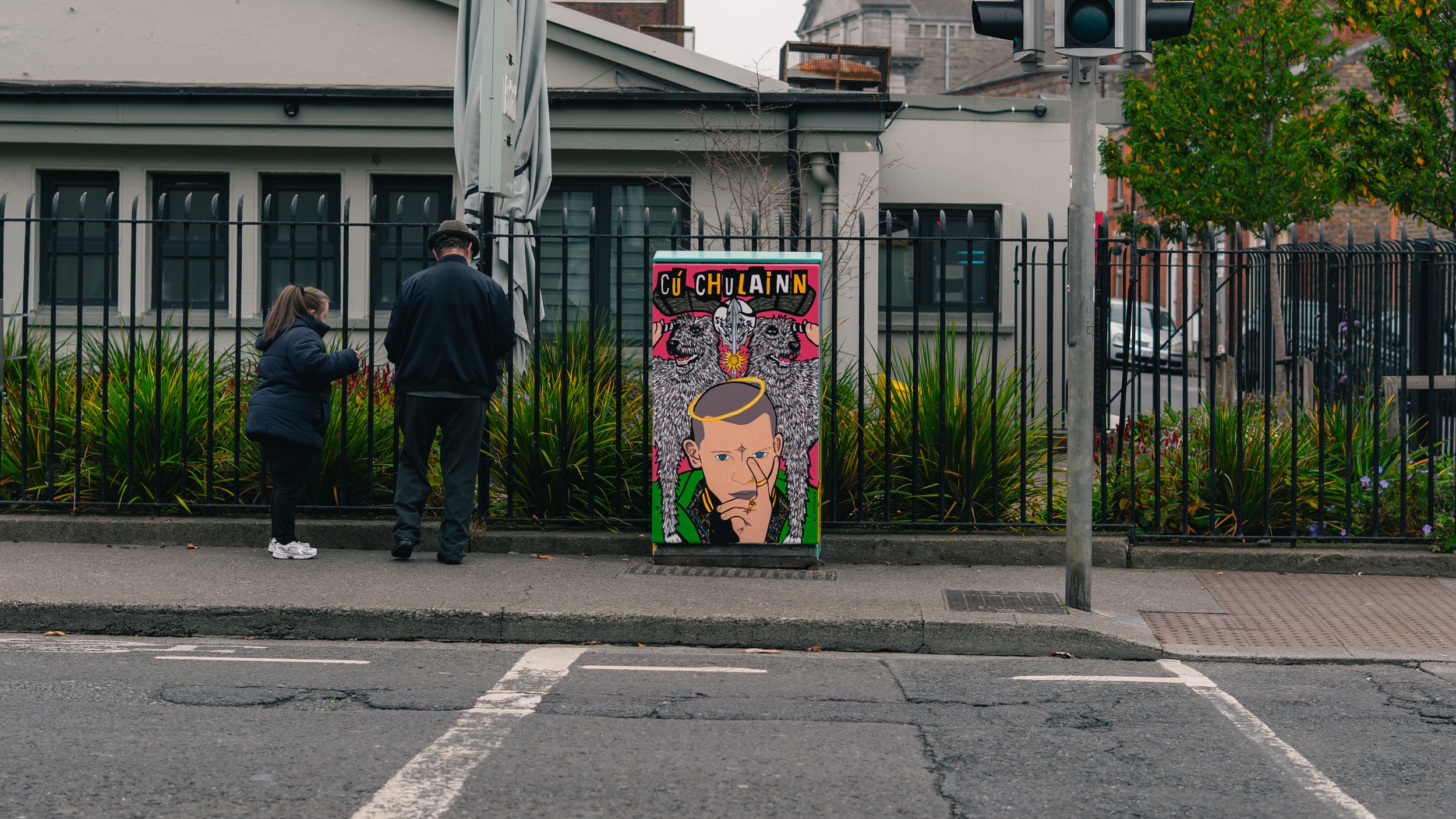 People standing on sidewalk near colorful street art sign with two dogs, a woman and man with their backs turned, and a fenced garden with green plants in an urban area.