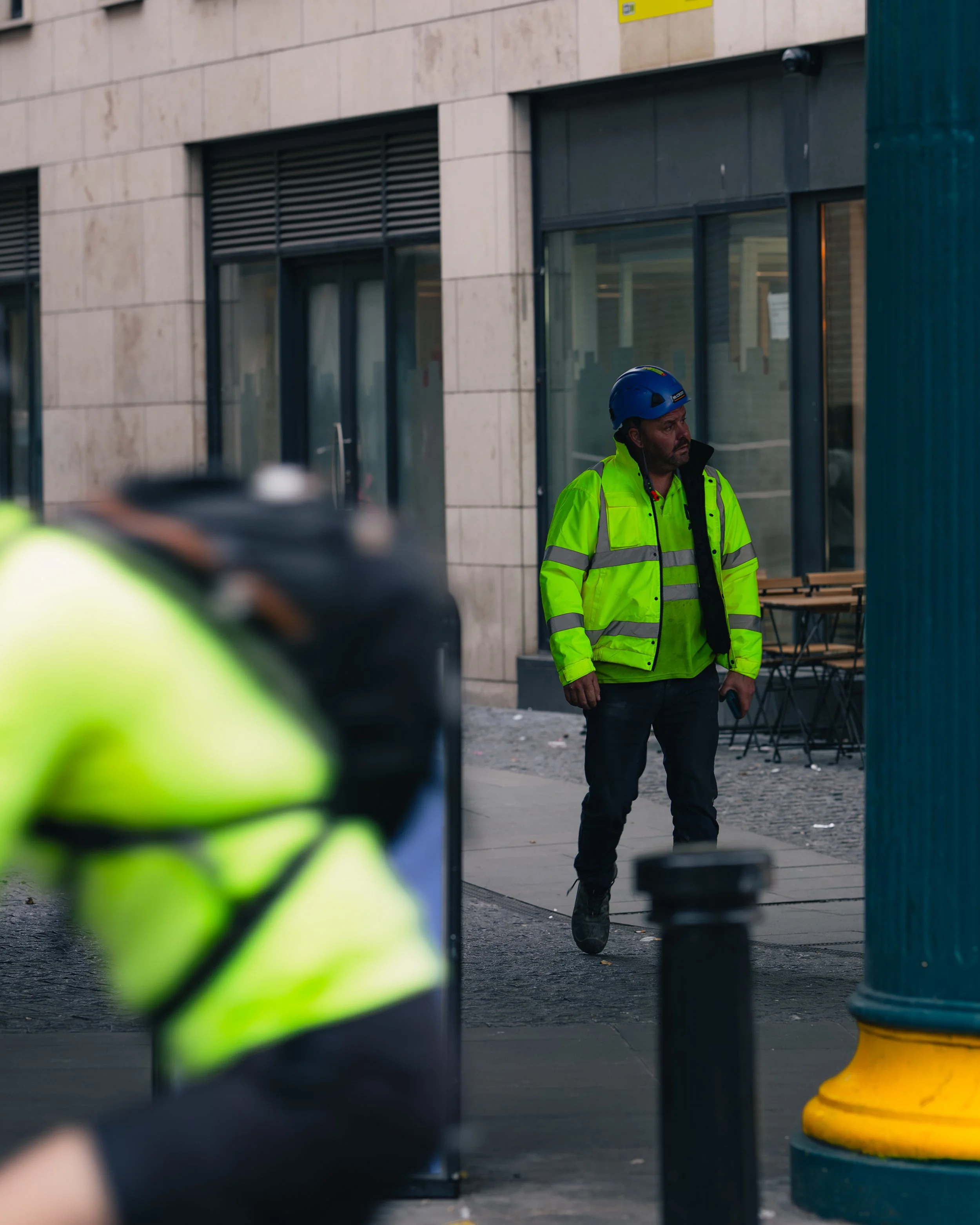 Man in a yellow reflective jacket and blue safety helmet standing on an urban sidewalk near a building.