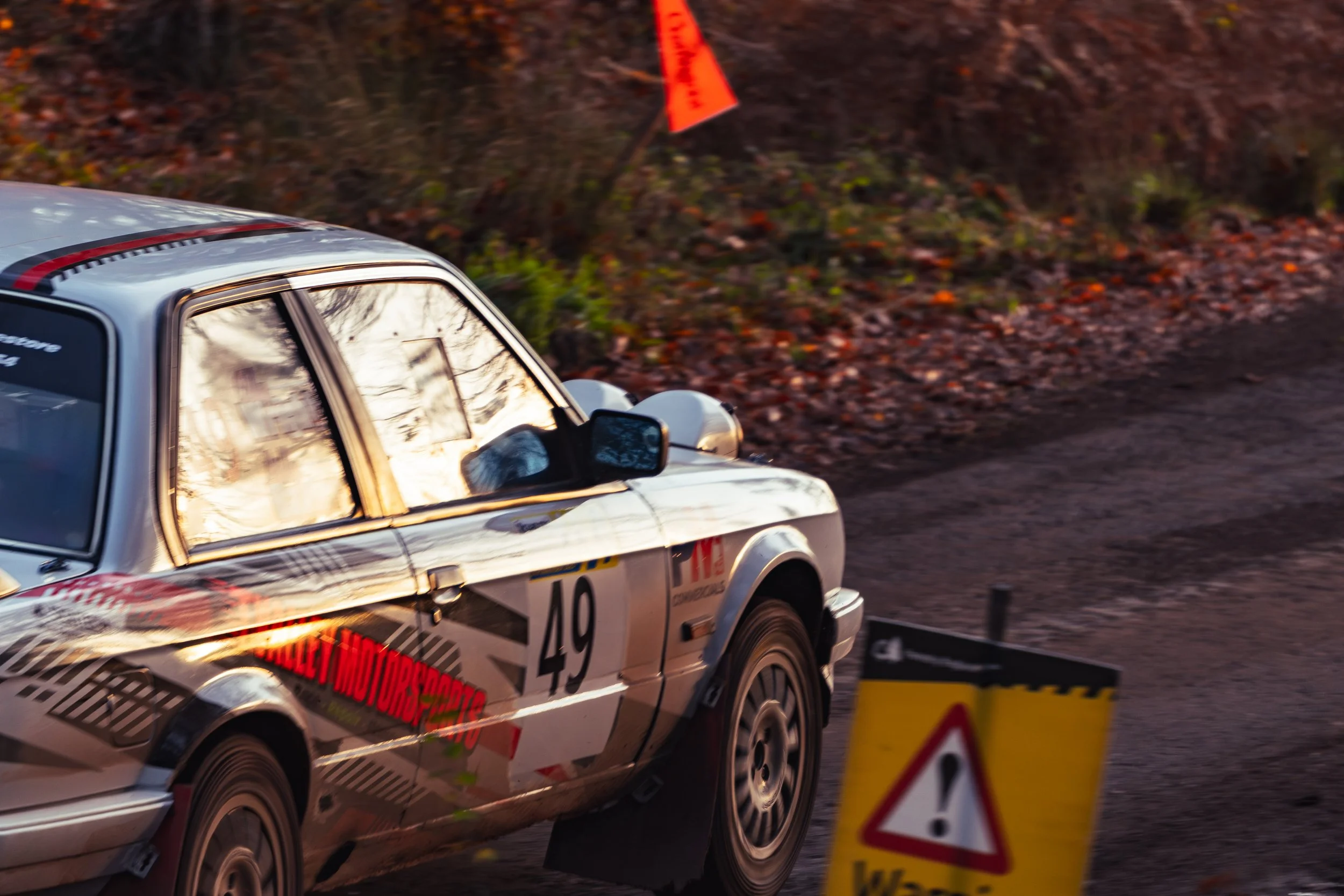 A silver race car with the number 49, racing decals, and a driver inside, is on a dirt track during a race. The scene is captured at dusk with a warning sign with an exclamation mark in the foreground. wye dean rally 