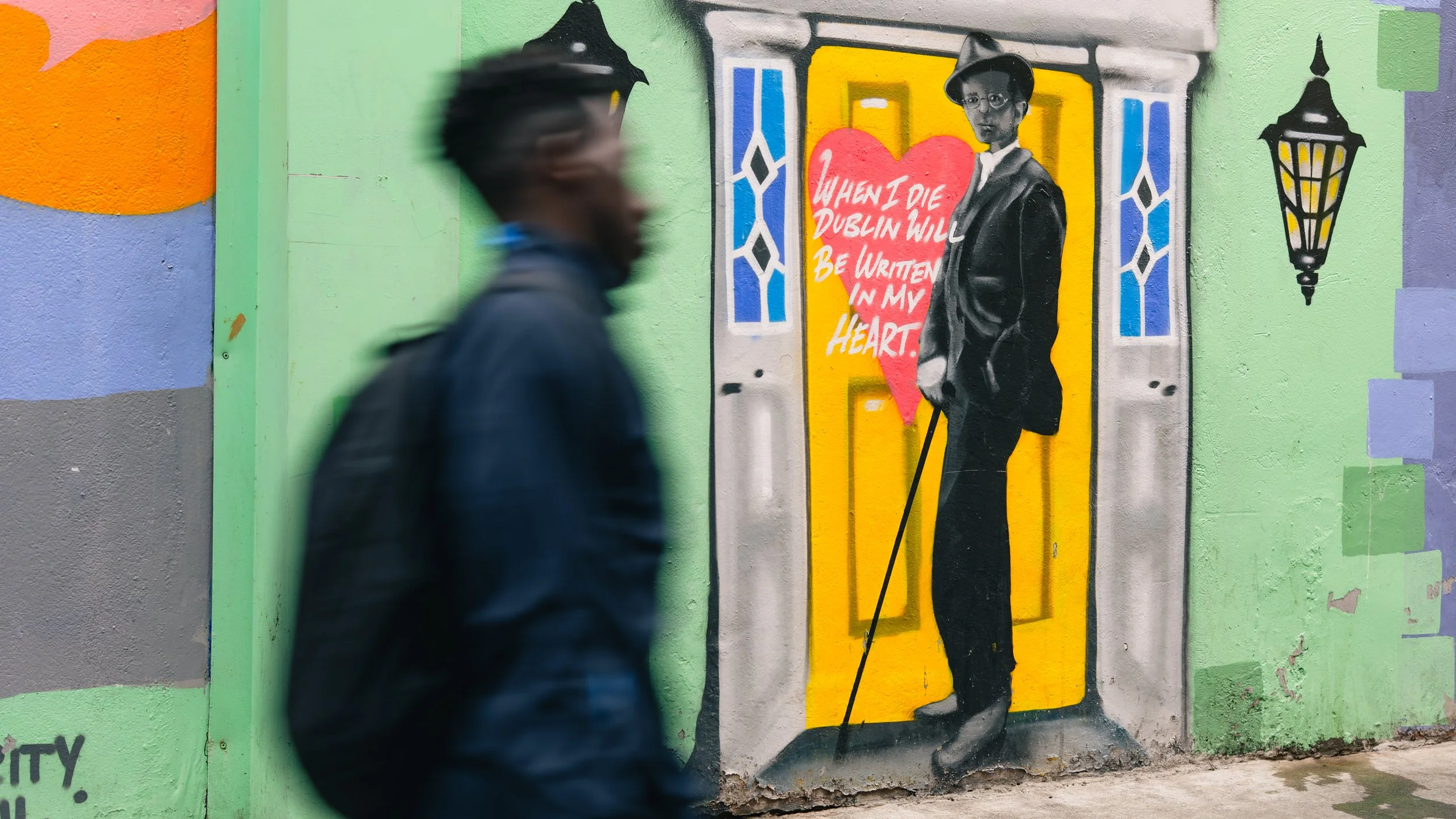 Street art mural of a woman in black and white standing in front of a yellow door with a red heart shape that has the words "When I die Dublin will be written in my heart." A man in dark clothing and glasses passes by in the foreground, appearing blu