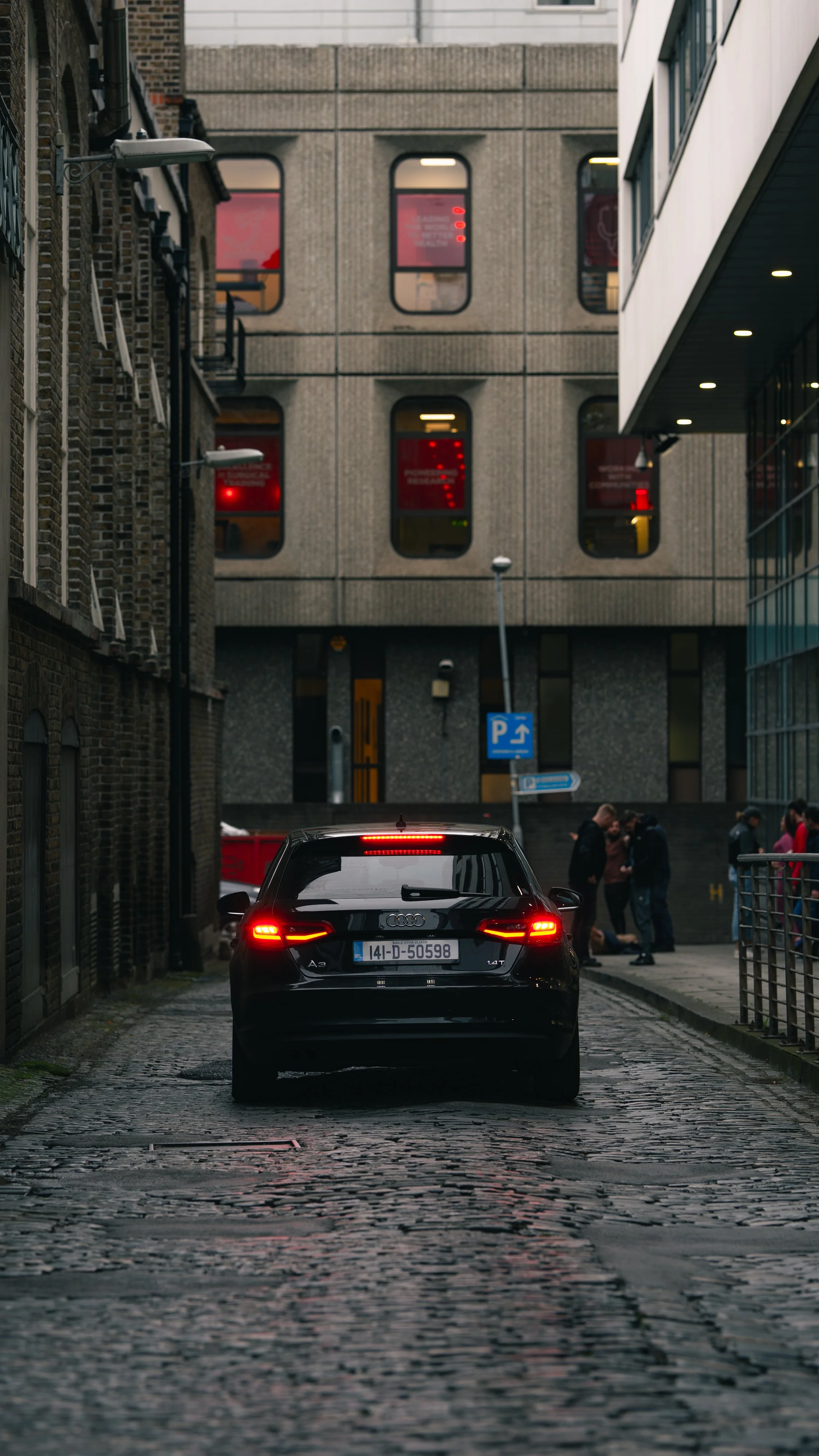 A black Audi A3 parked on a cobblestone street at dusk with an urban background, including modern and older buildings and a group of people on the sidewalk.