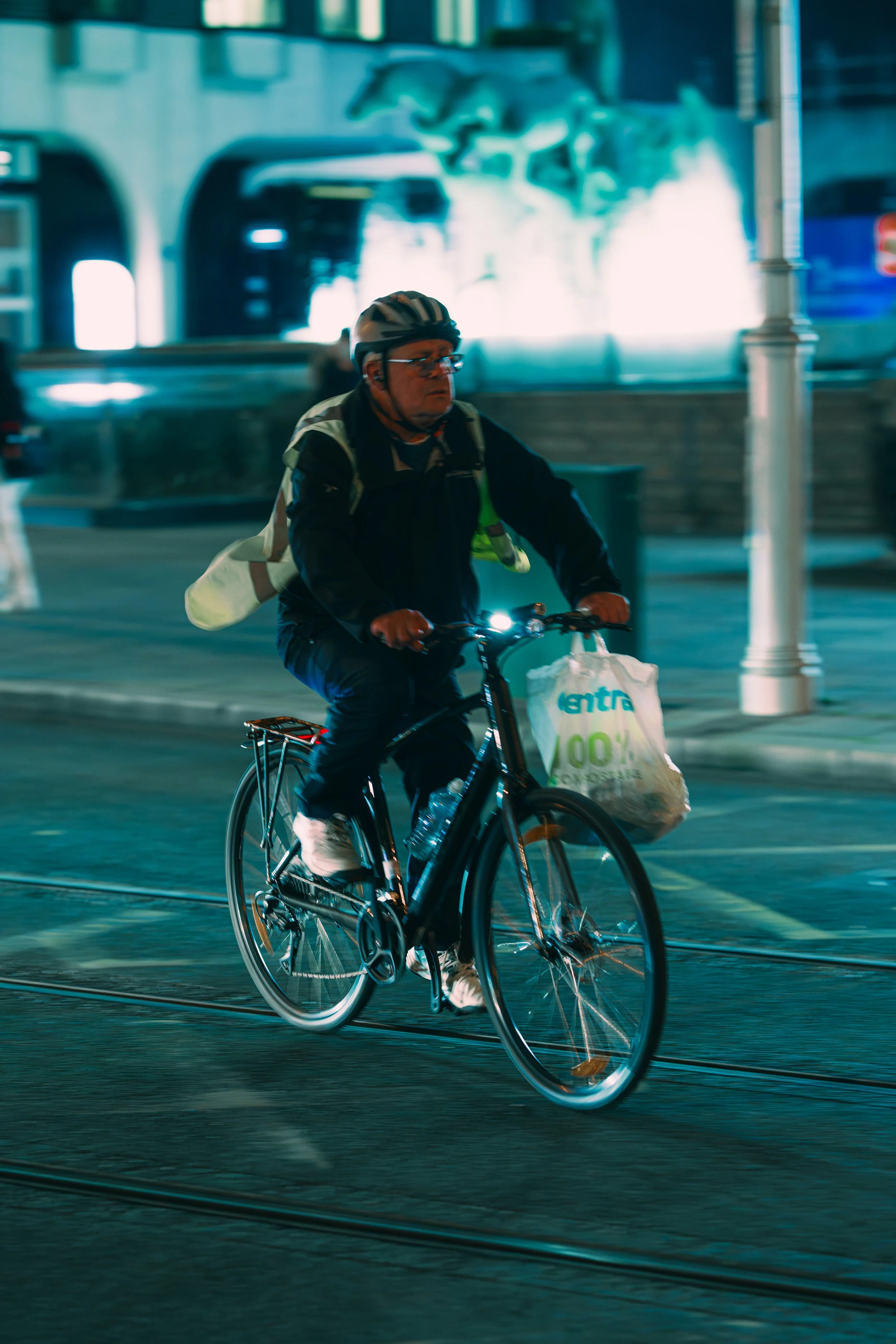 A man wearing a helmet and glasses riding a bicycle at night, carrying a plastic bag labeled 'entra' hanging from the front of the bike, with a city street and a painted mural in the background.