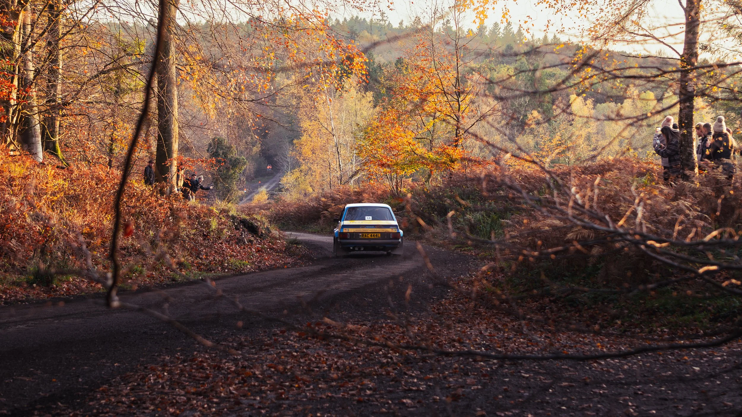A white rally car on a dirt road surrounded by autumn trees with orange and yellow leaves, with spectators standing along the hillside. wye dean rally 