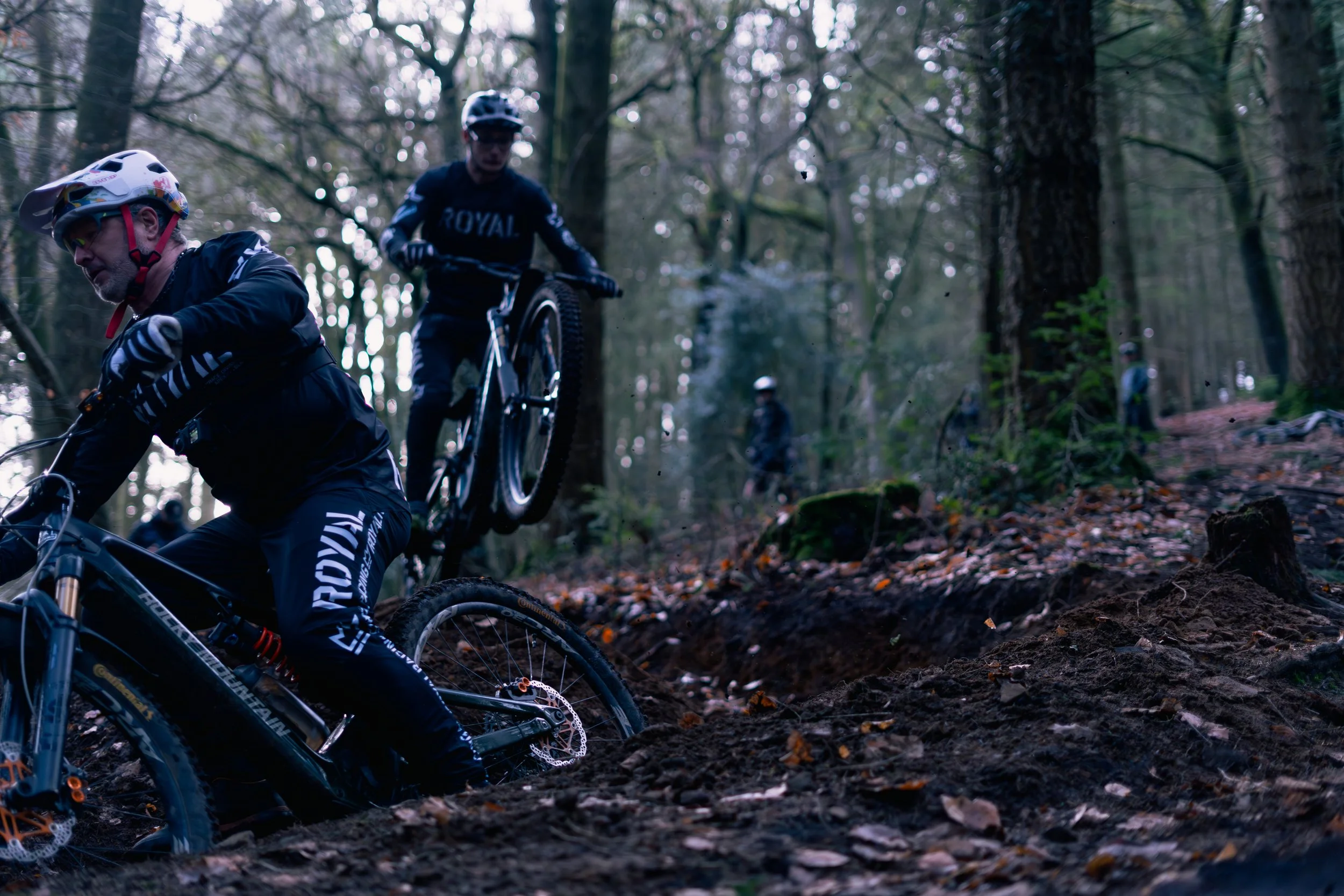 Three mountain bikers riding on a dirt trail in a forest with trees and fallen leaves.