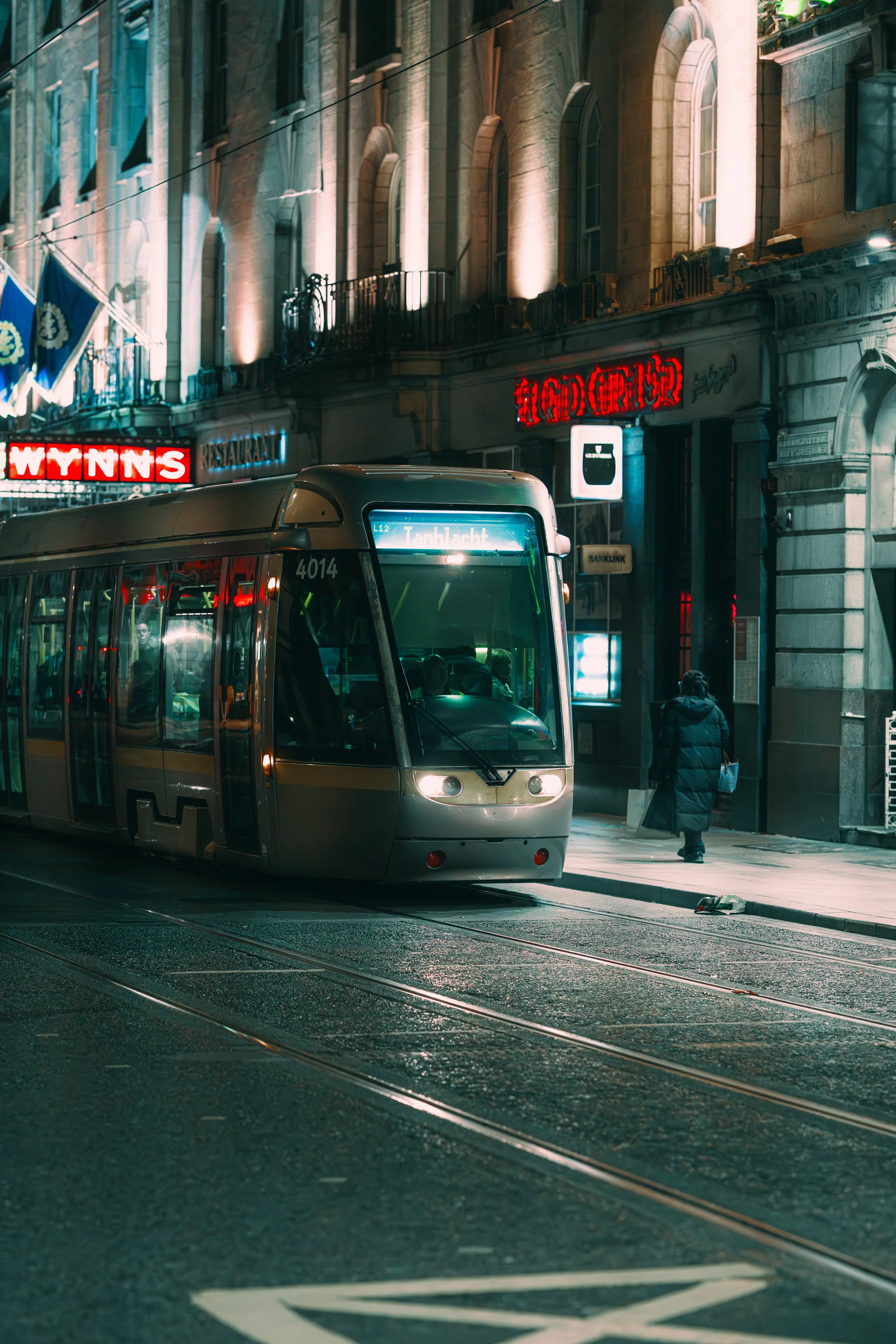 A modern tram on city tracks at night, with illuminated signs and buildings in the background, including a neon sign that says 'WYNNS' and another that says 'HOGER'. A person in a dark coat walks on the sidewalk nearby.