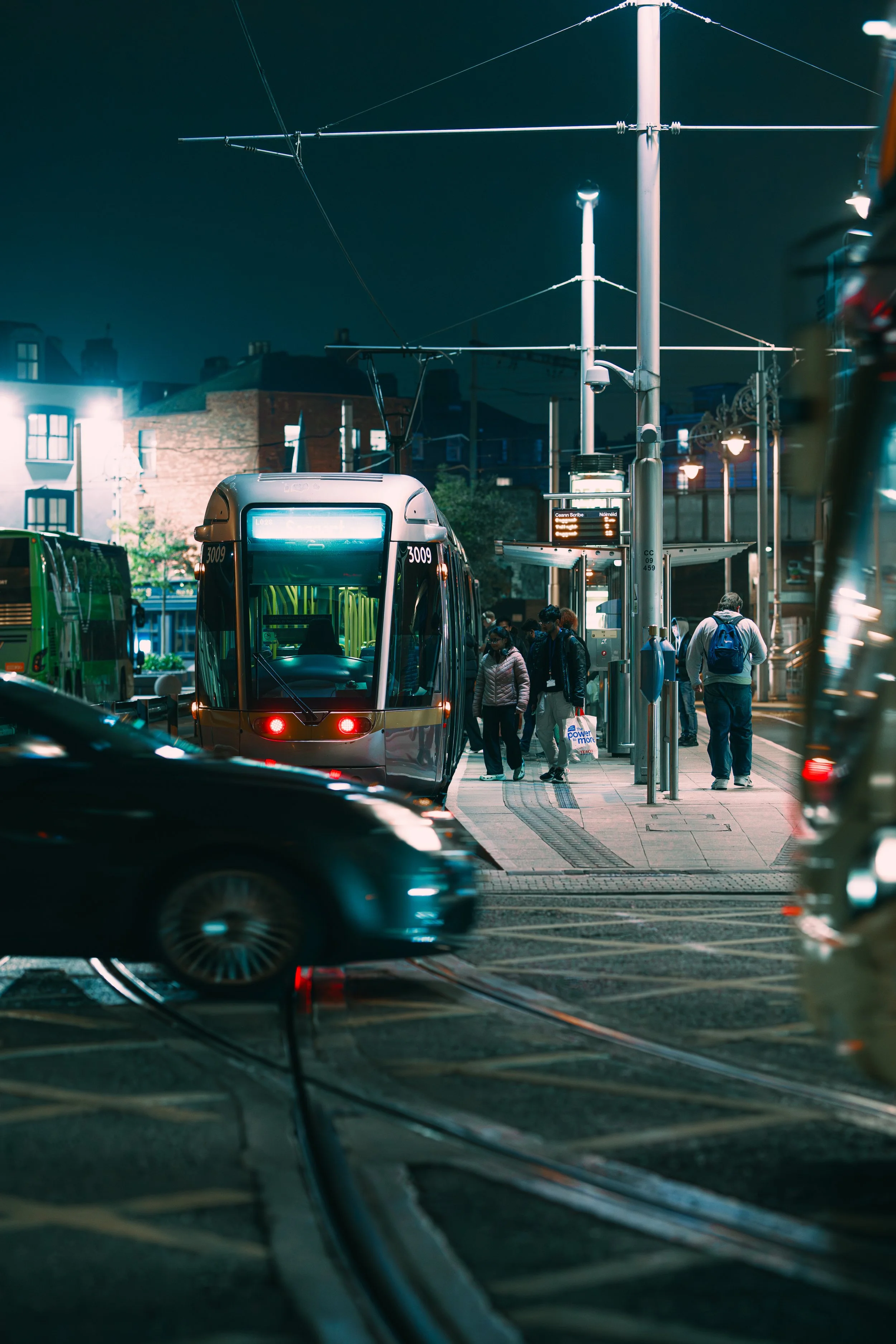 A city street at night with a modern tram arriving at a stop, pedestrians waiting, and cars passing by.