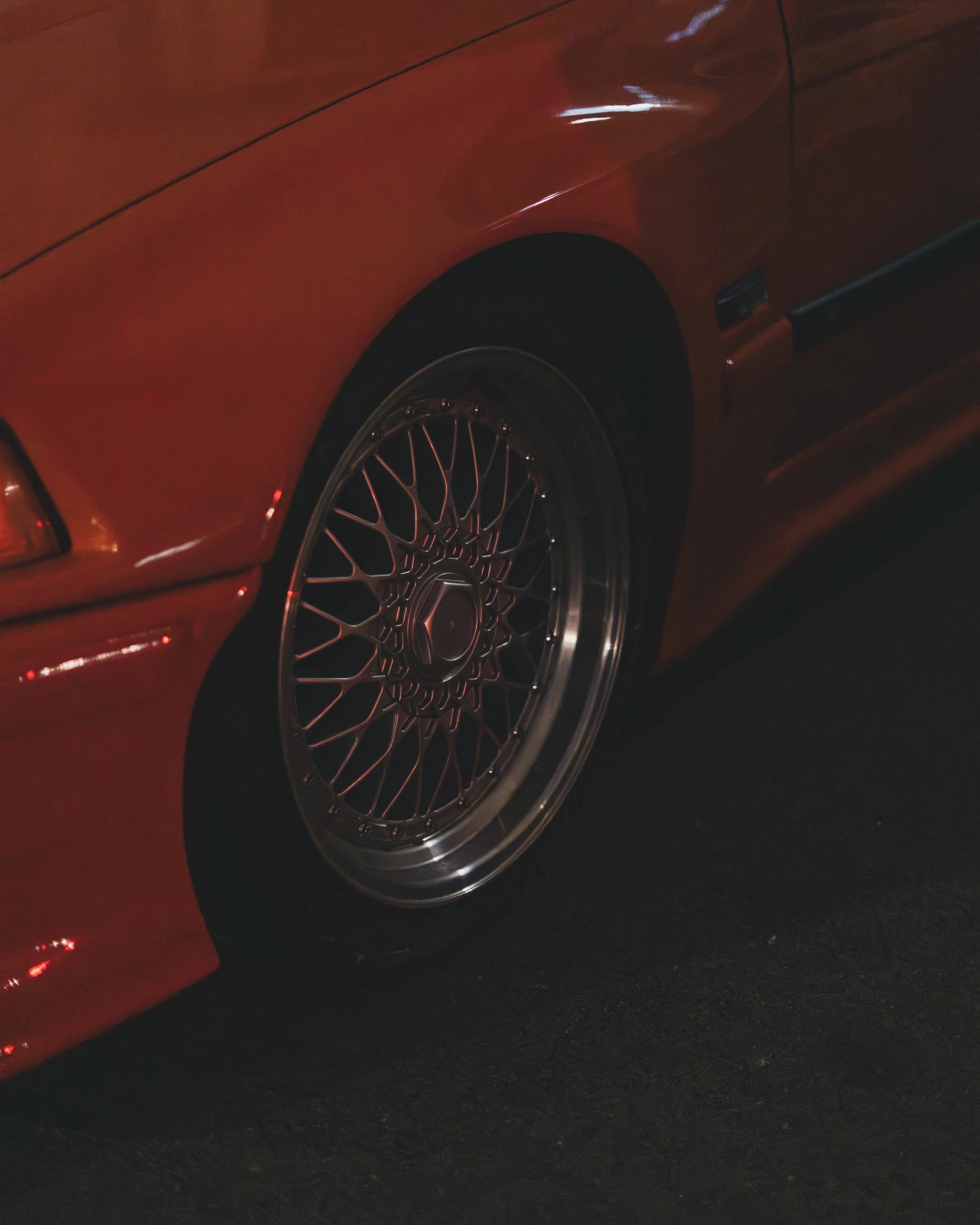 Close-up of a red sports car with a custom wire wheel rim on the front tire, parked on a dark surface.