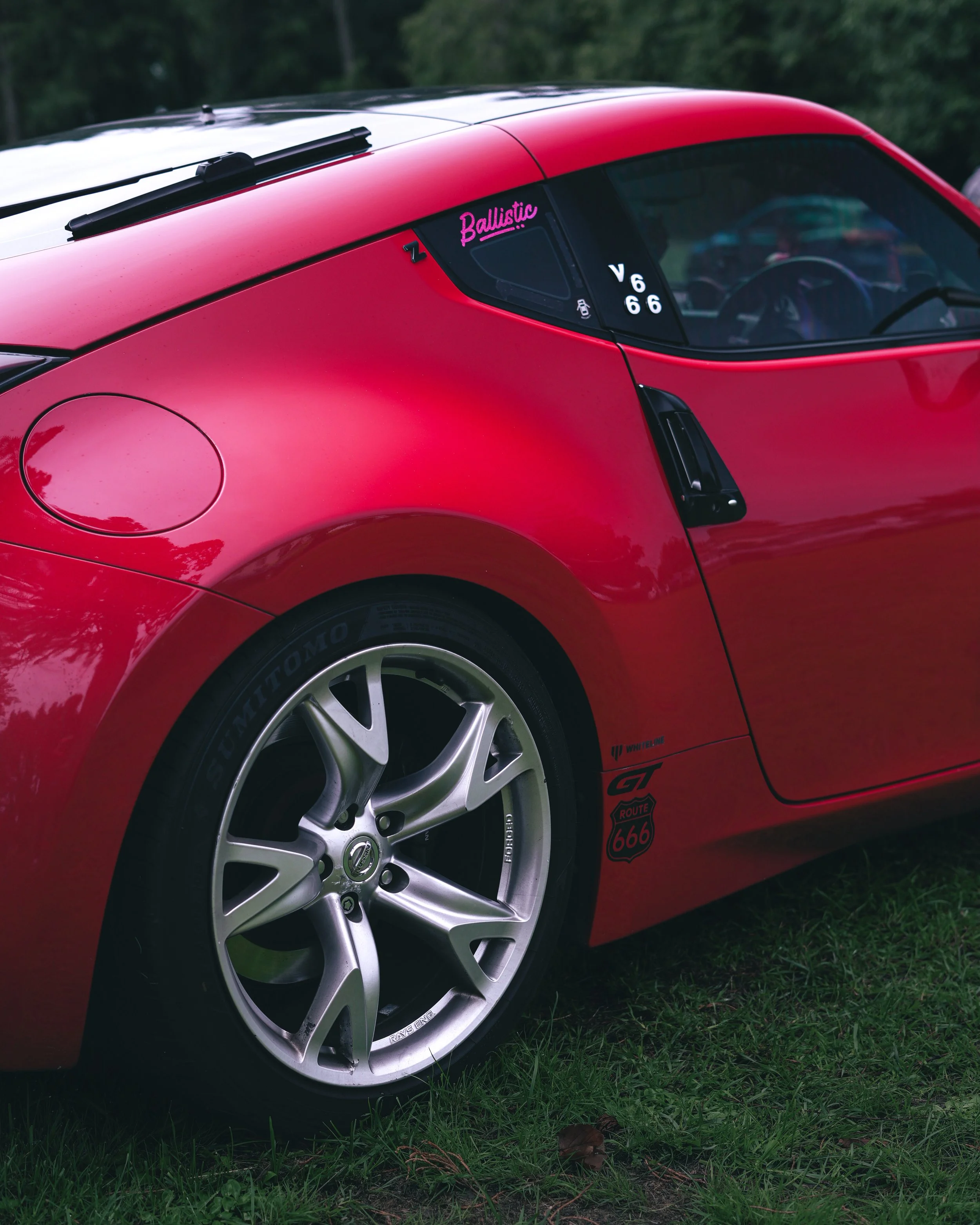 Close-up of a red  Nissan z370 sports car parked on grass, showing the rear wheel, side window with stickers, and part of the body with racing decals.