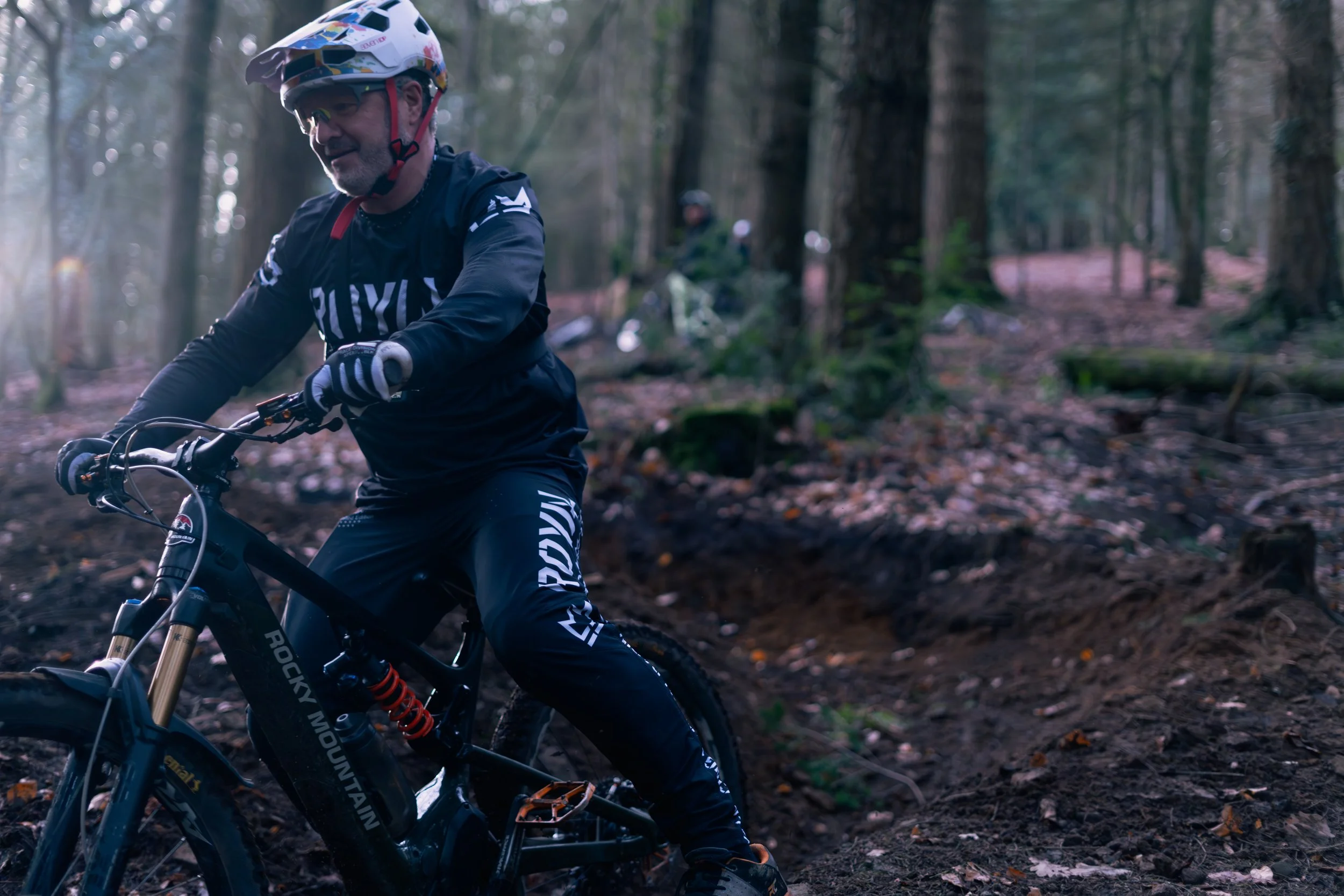 A man mountain biking on a dirt trail through a forest during daylight, wearing a helmet and sports gear.