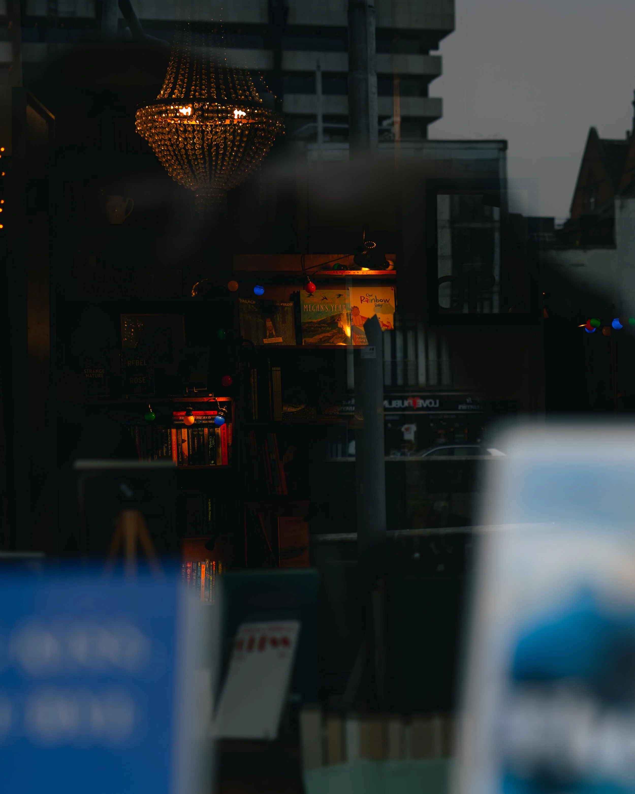 Very dark image of a bookstore or library interior with a warm chandelier and colorful string lights, visible bookshelves, and some books on display.