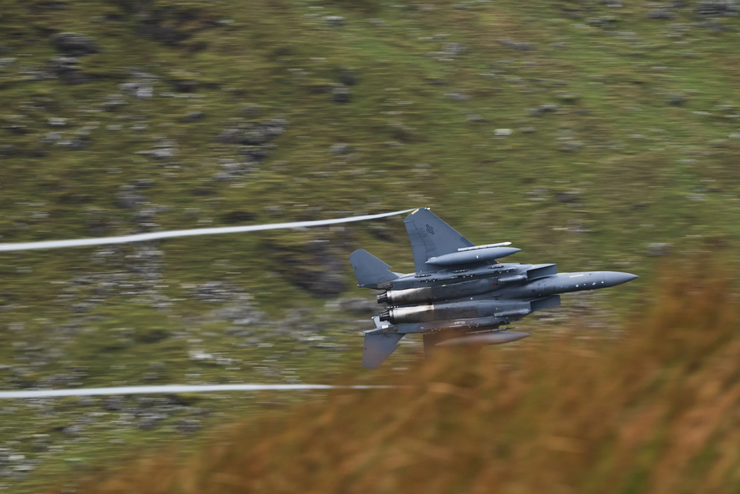 A fighter jet flying low over a grassy landscape, leaving a vapor trail behind.