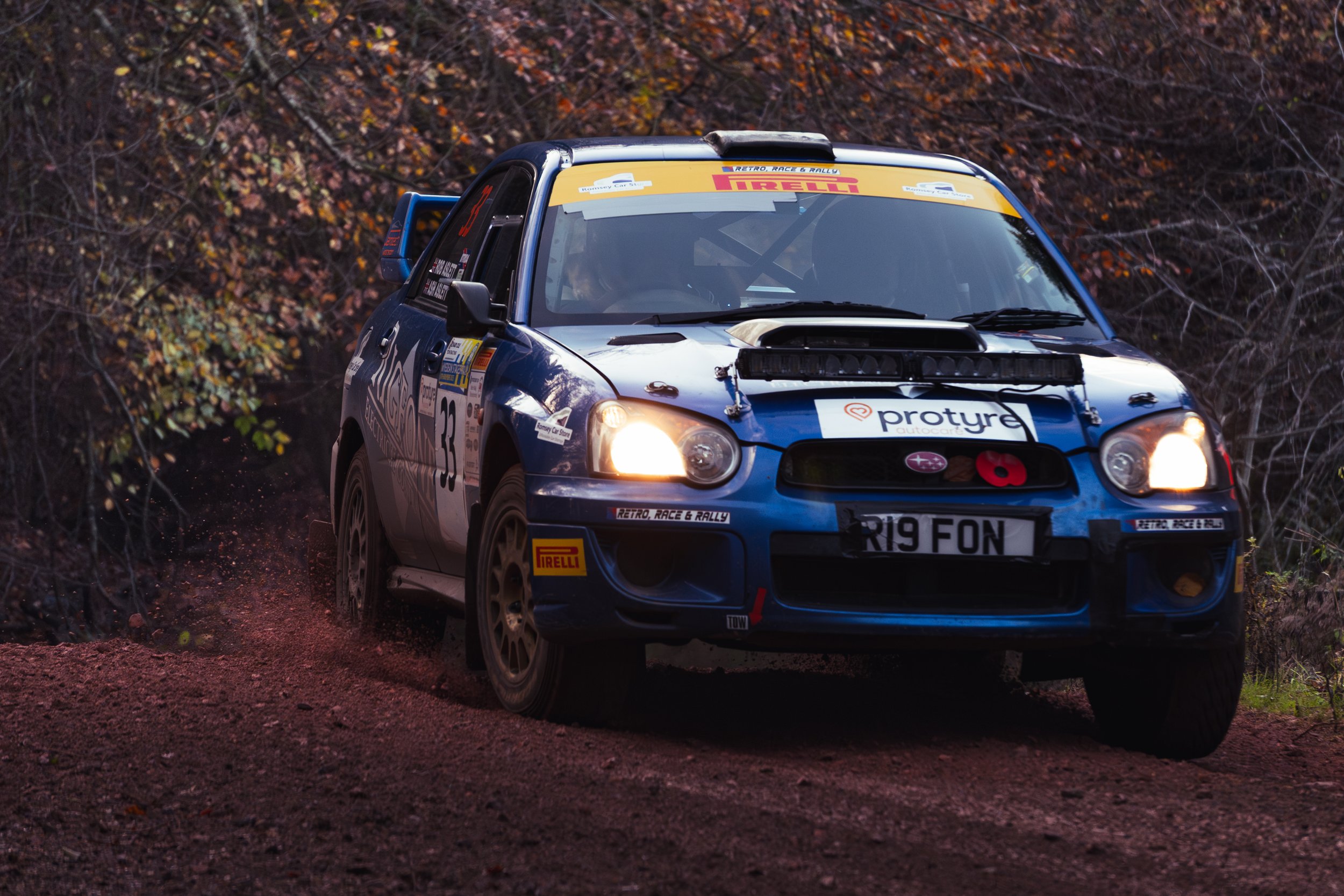A blue rally car with headlights on driving on a dirt trail surrounded by trees with autumn leaves. wye dean rally 