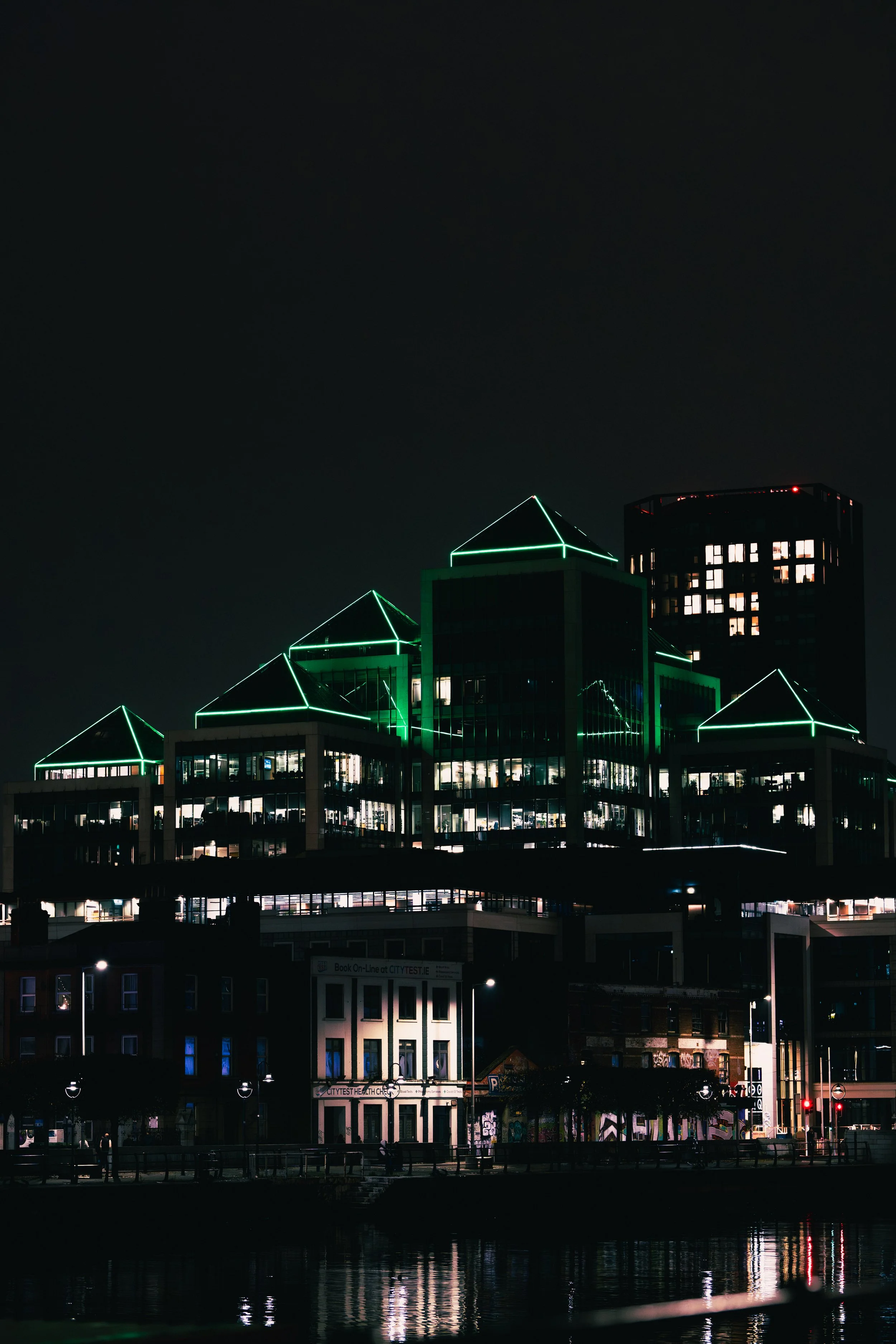 Nighttime cityscape featuring a modern glass building with green neon outlines on its rooftops, reflecting in water below.
