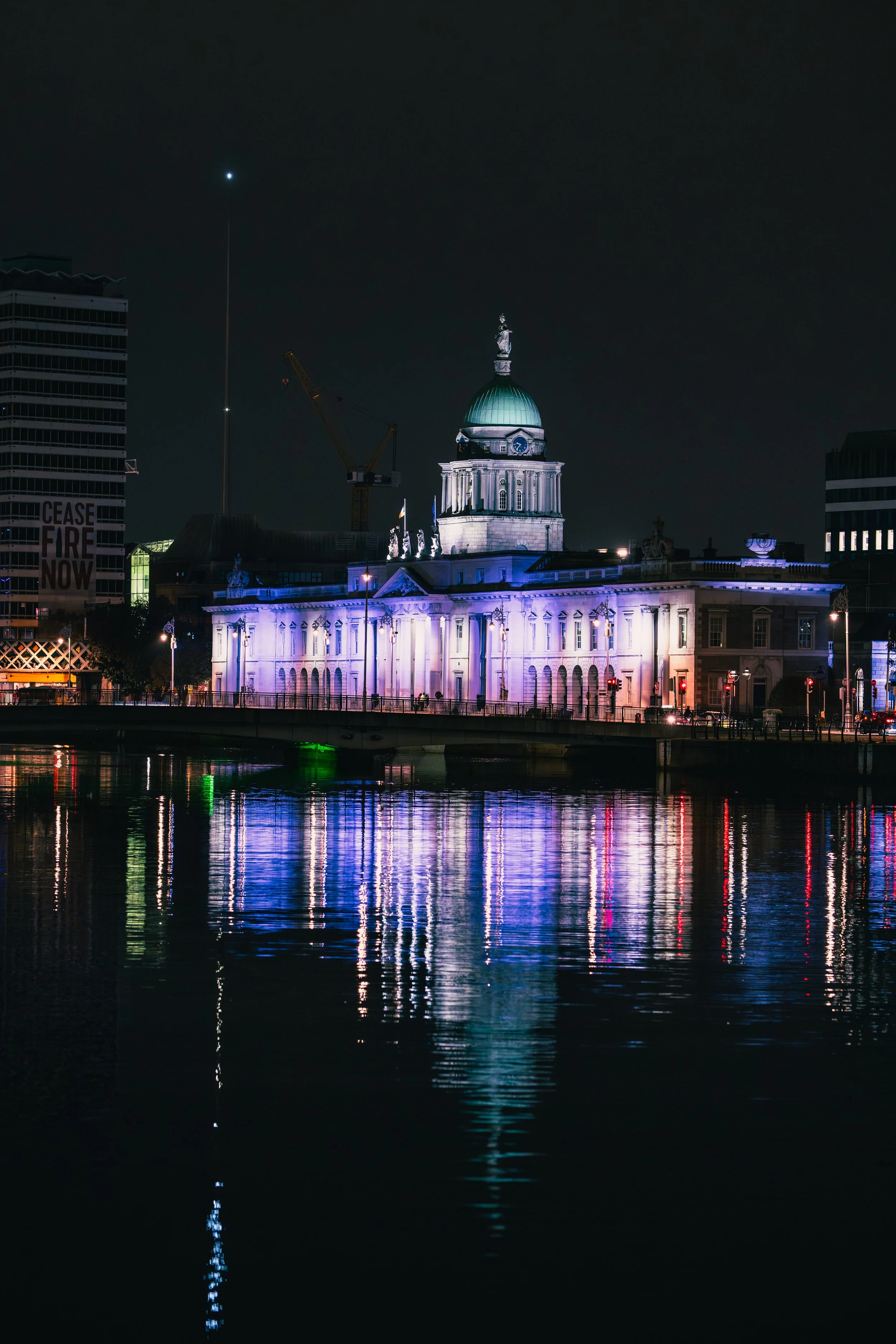 Nighttime view of a historic government building illuminated with purple and white lights, reflecting on the water in front of it.