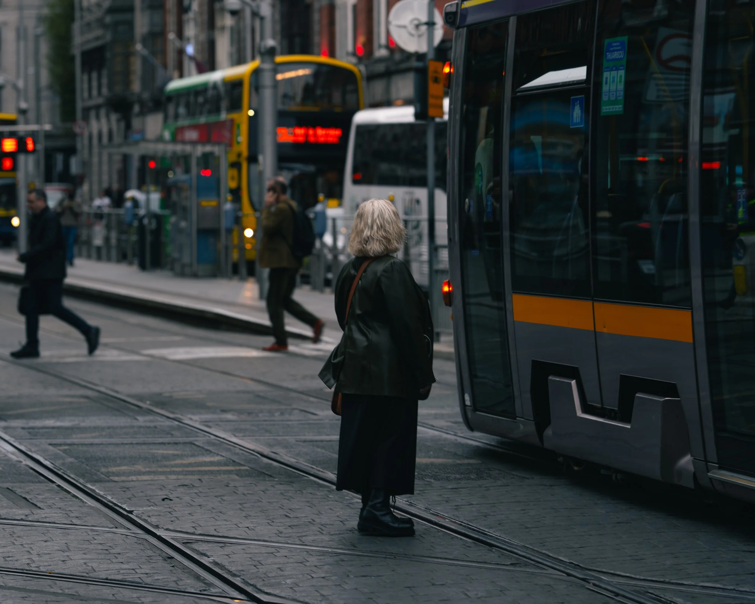 A woman with gray hair standing at a city tram stop, waiting to board a modern tram on a busy street with buses and pedestrians.