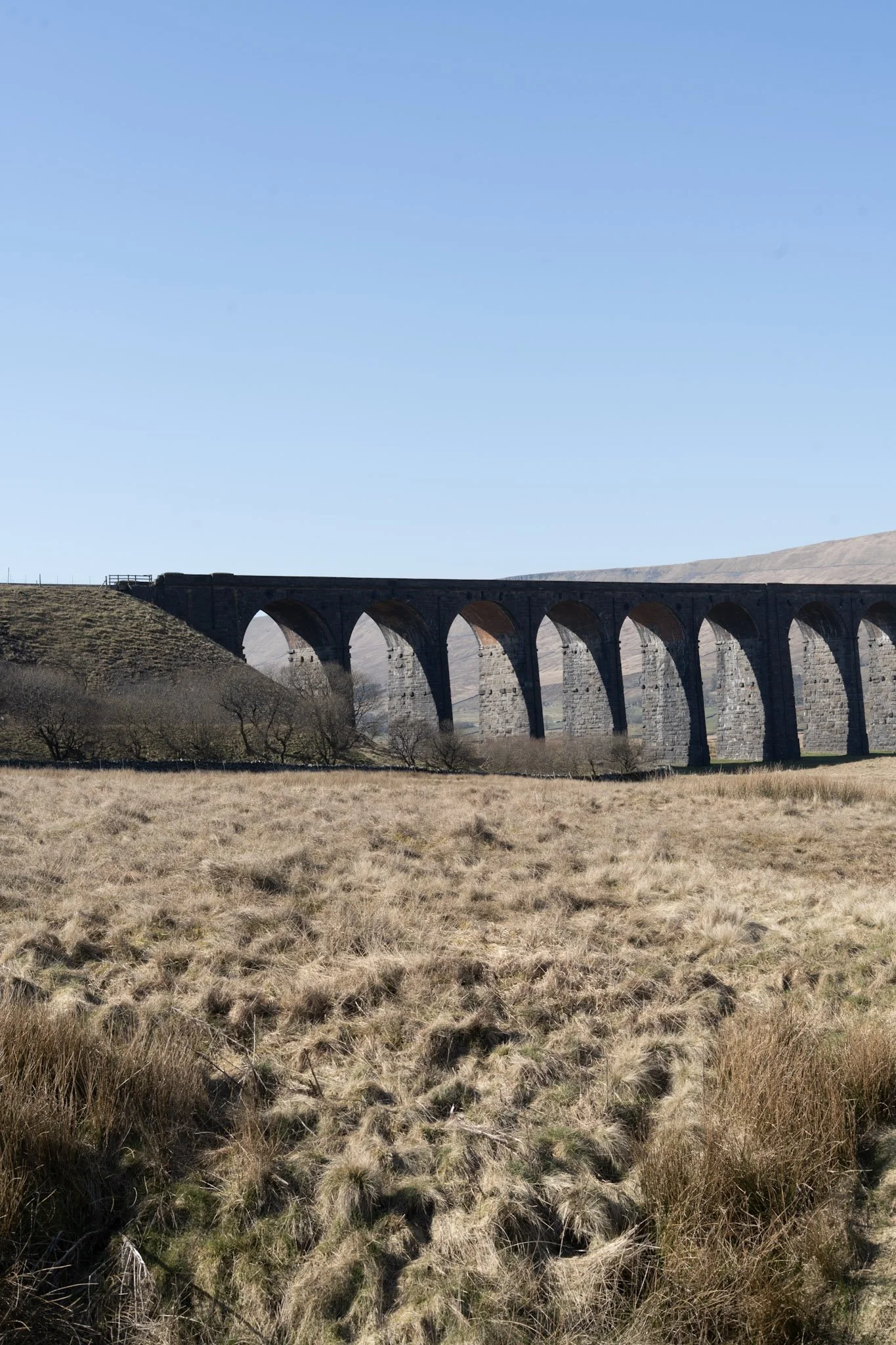 A stone arch bridge extending across a rural landscape with dry grass and sparse trees, under a clear blue sky.
