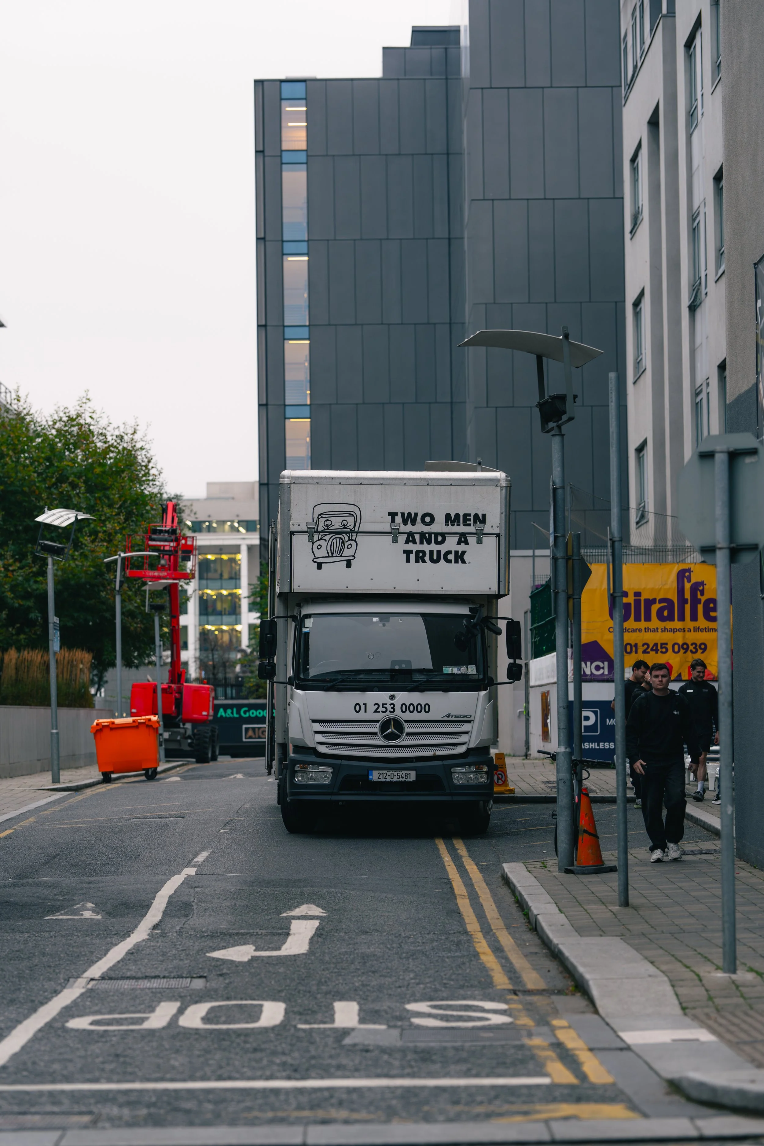 A city street with a white truck displaying a sign that reads 'Two Men and a Truck' parked in a lane with a 'STOP' painted on the road. There are pedestrians on the sidewalk, and modern buildings in the background.