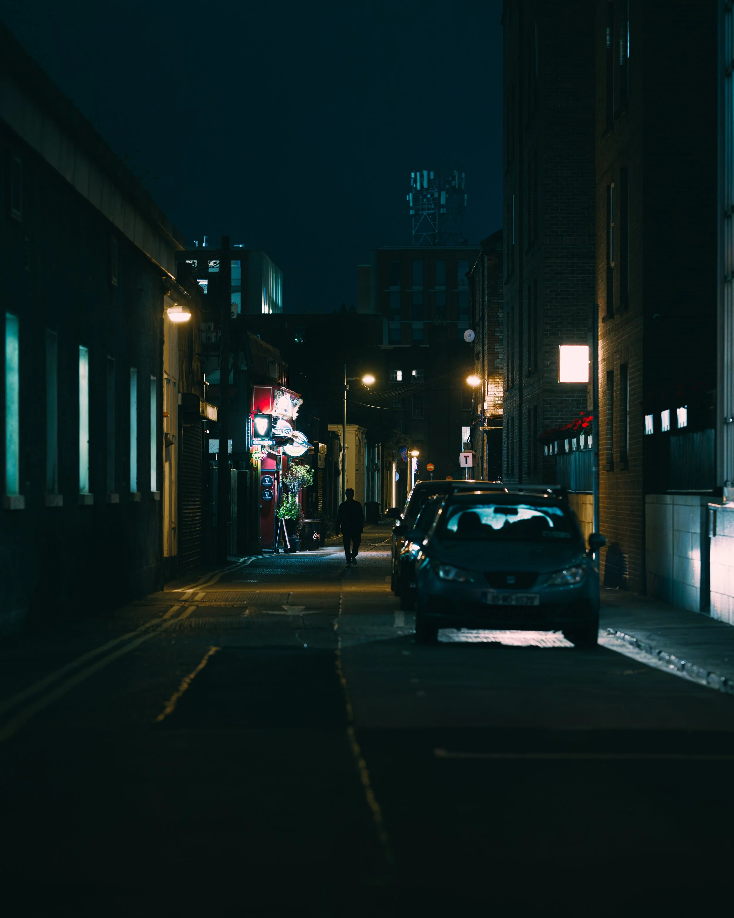 Nighttime urban street scene with parked cars, a solitary walking figure, illuminated signs, and buildings with lit windows.