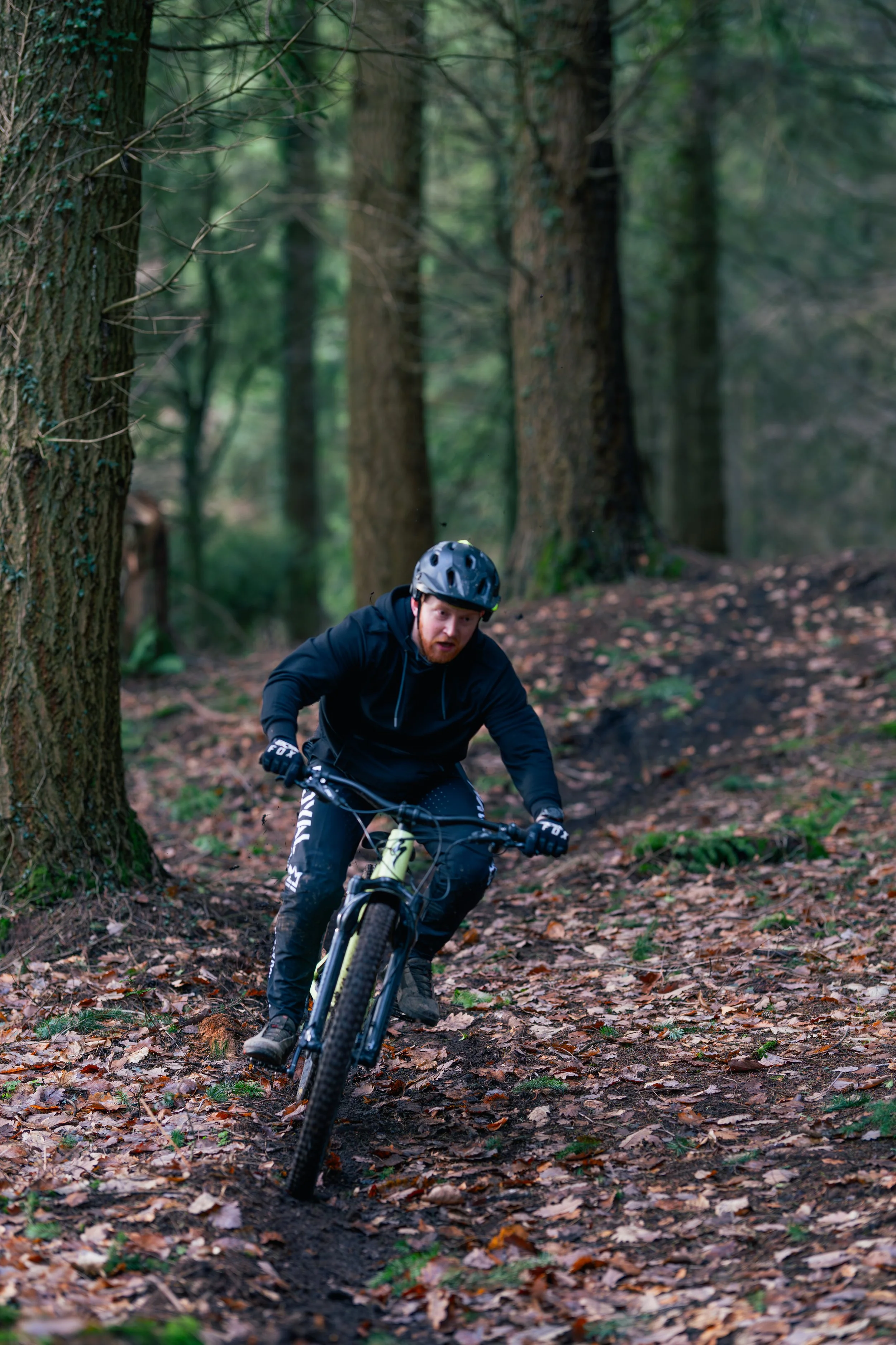 A man in a black jacket and helmet riding a mountain bike on a trail through a forest with tall trees and fallen leaves.