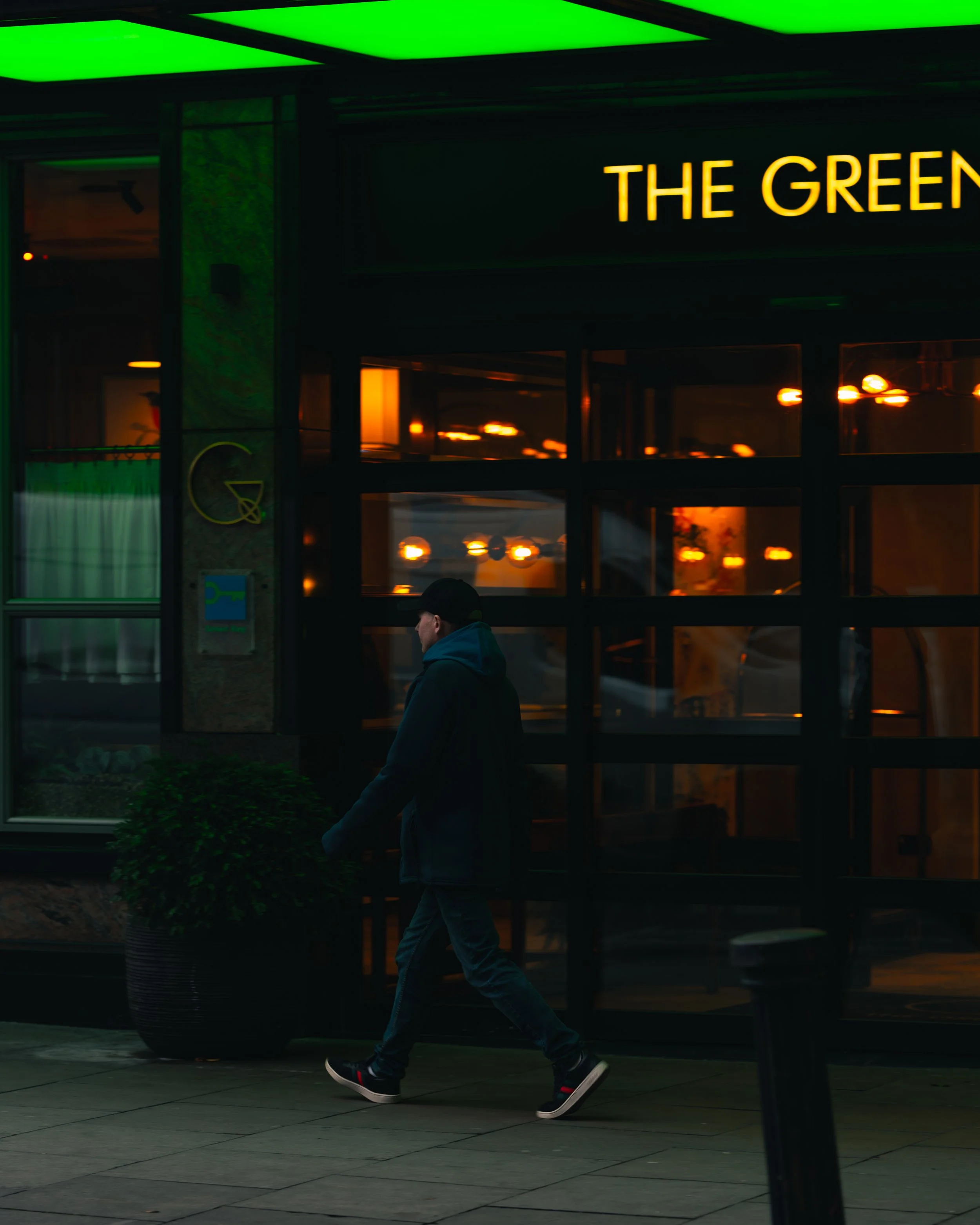 A person walking past a restaurant with a lit sign reading 'The Green' at night, reflections of warm lights visible through the windows.