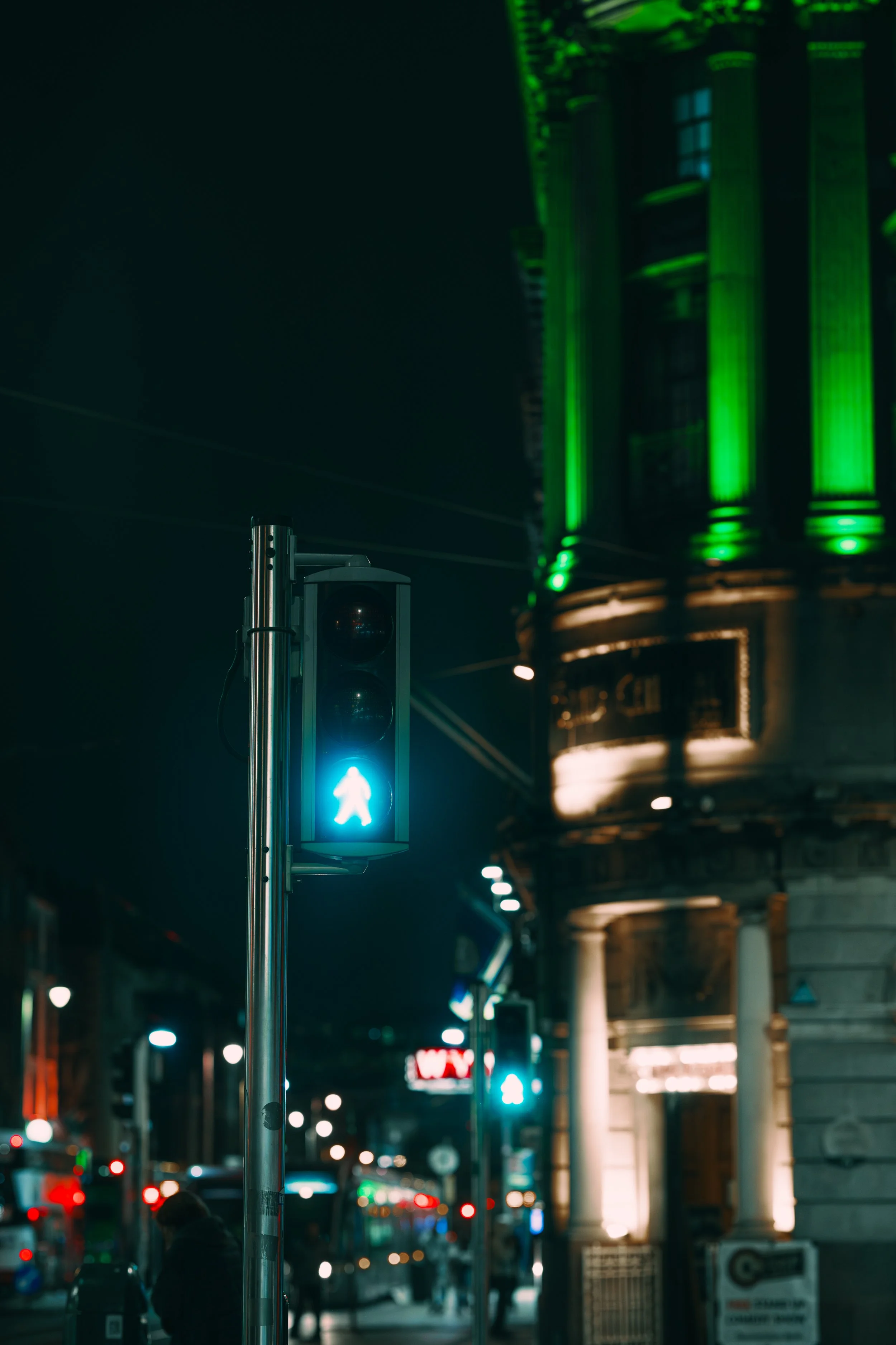 A city street at night featuring a green pedestrian traffic light, a historic building with green and white lighting, and blurred city lights and people in the background.