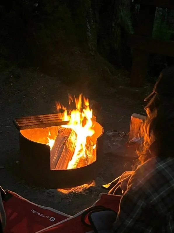 A wood-burning fire within a BC Parks campground fire pit is lit. The fire illuminates the shoulder of a person with long brown hair in the bottom right corner. Over the image is a location description with text reading: French Beach