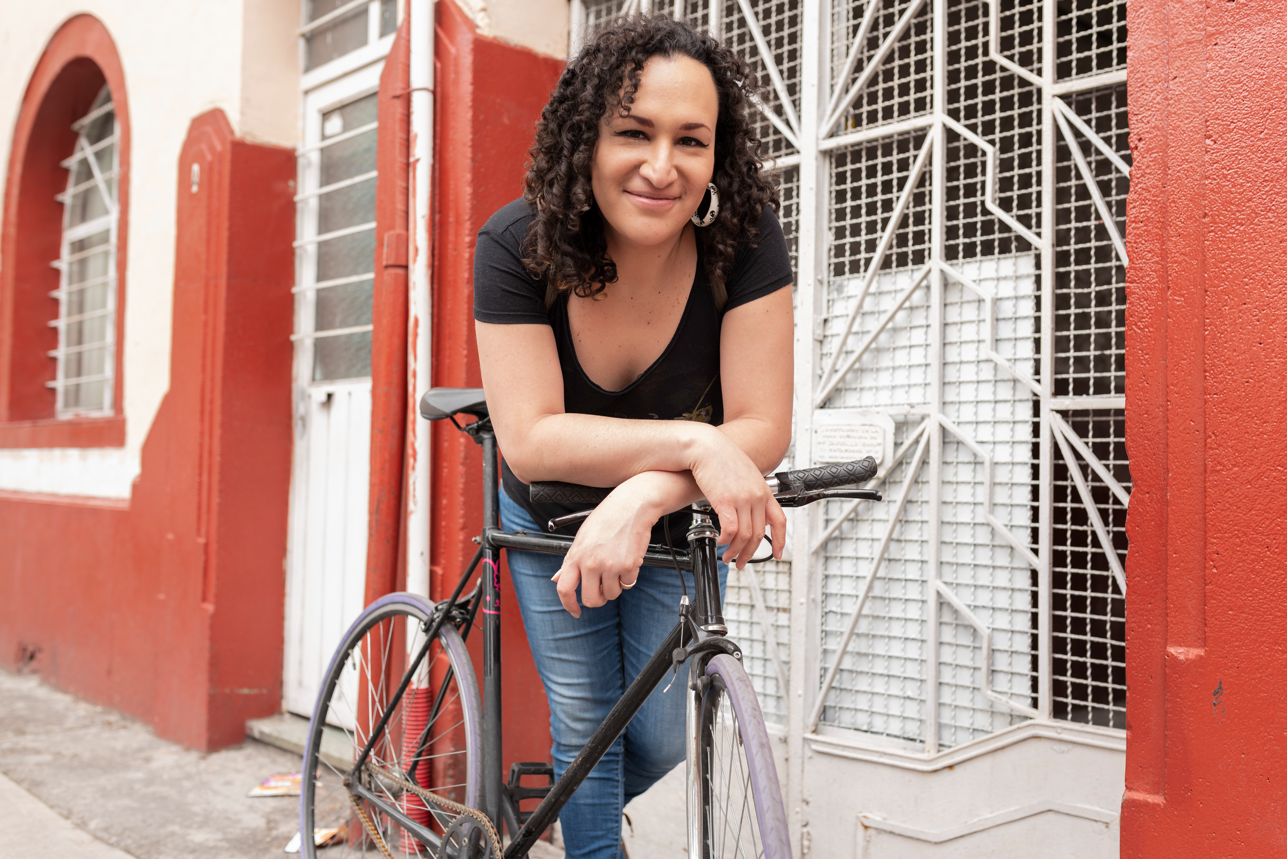 Person smiling at the camera resting their forearms. They have long curly brown hair, winged black eyeliner, a v-neck black top, and a hoop earring.