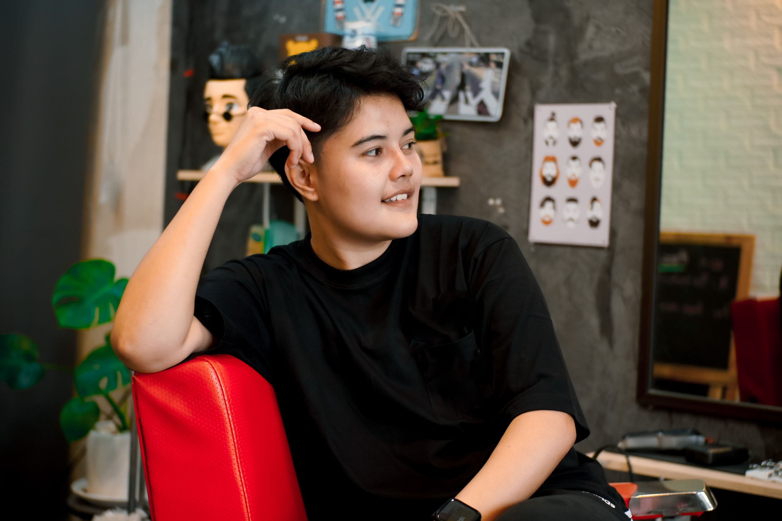 A person with short brown hair sits in a bright red chair looking off-camera to the right. The person has their right hand rested on their head. They have a neutral expression