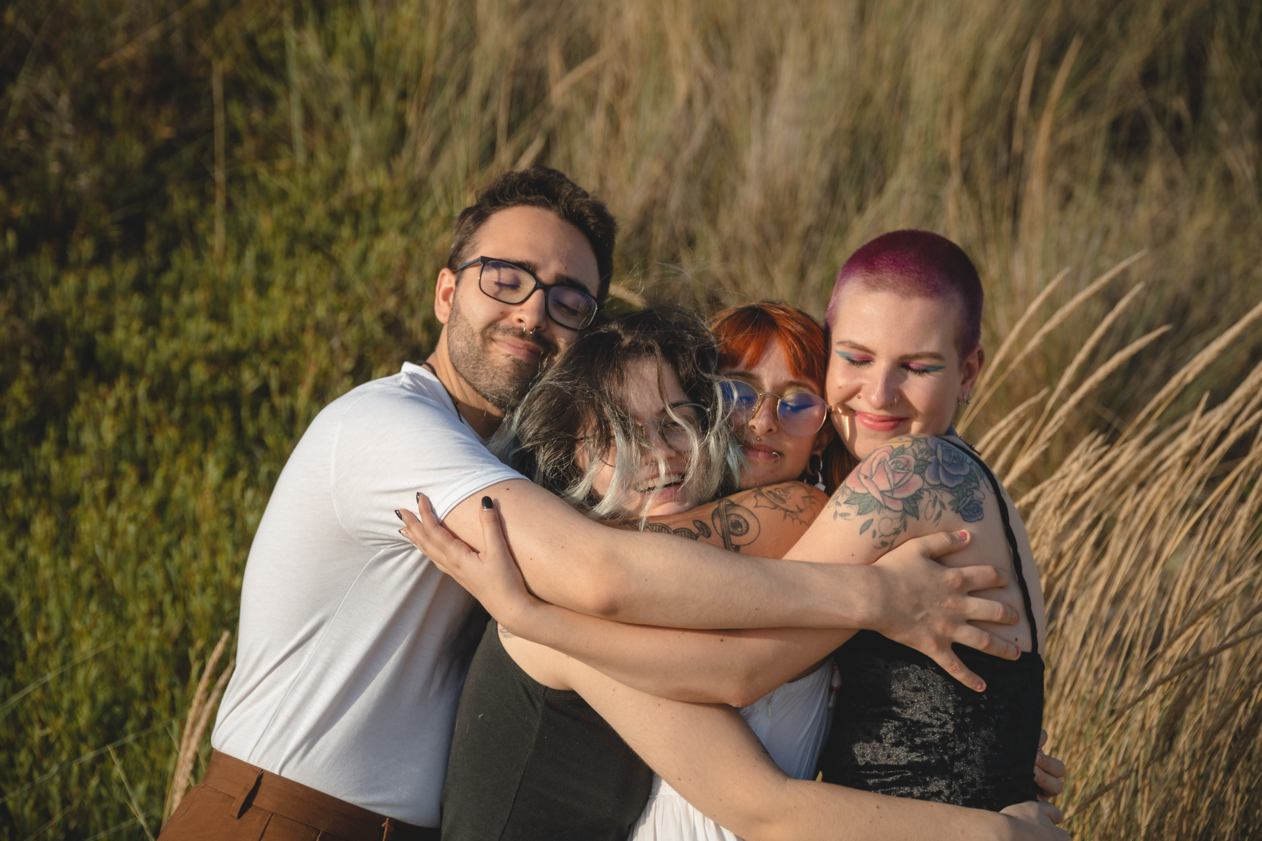 Four people face the camera smiling with eyes closed holding each other. From left to right: person 1 glasses, white shirt, septum piercing; person 2 long hair, glasses, black tank top; person 3 tattooed arm, orange hair, bangs;person 4 pink buzz cut