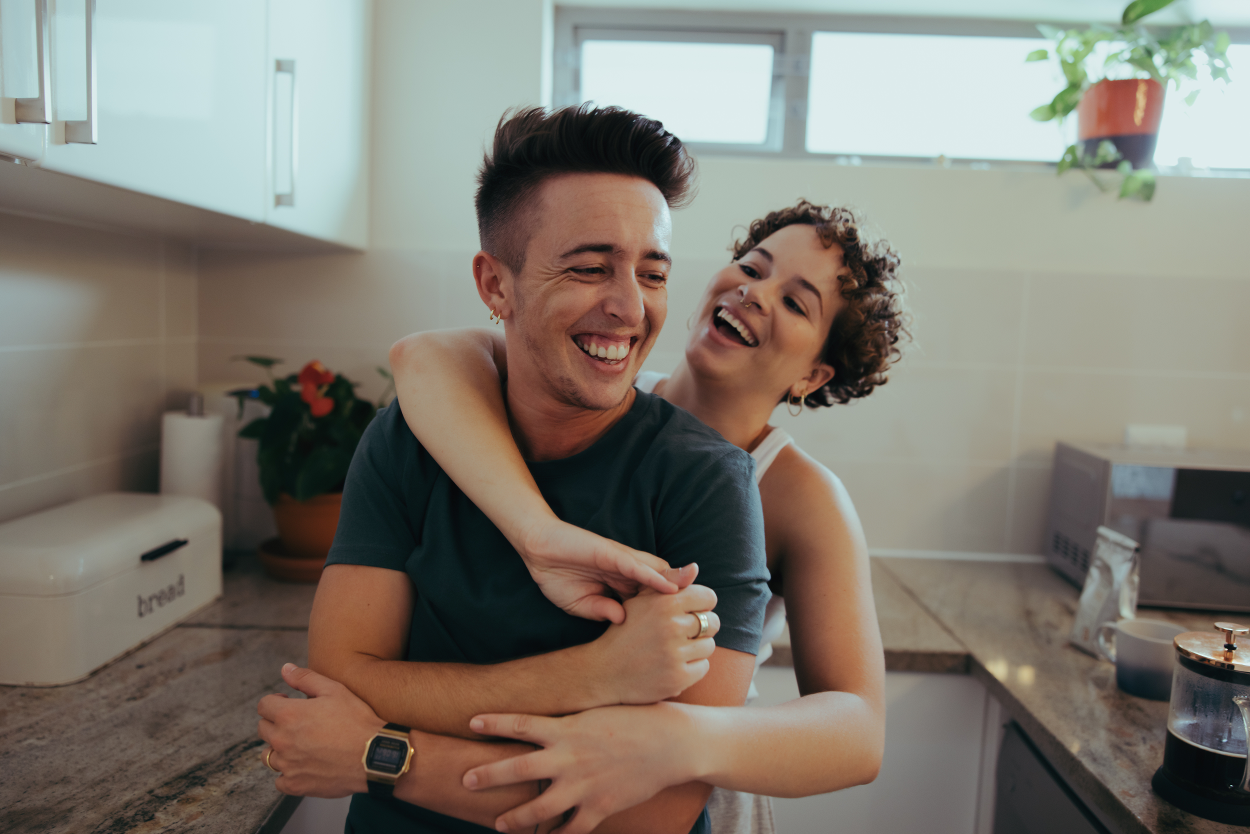 Two people standing in a kitchen. Both are smiling and the person in the back has their arms wrapped around the other, they have short curly hair, earrings, and a septum piercing. The person being hugged has short brown hair with a side fade