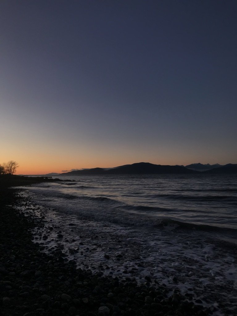 A sunset-lit sky with the North Shore mountains in the background and the pacific ocean in the foreground. Over the image is a location description with text reading: Spanish Banks beach
